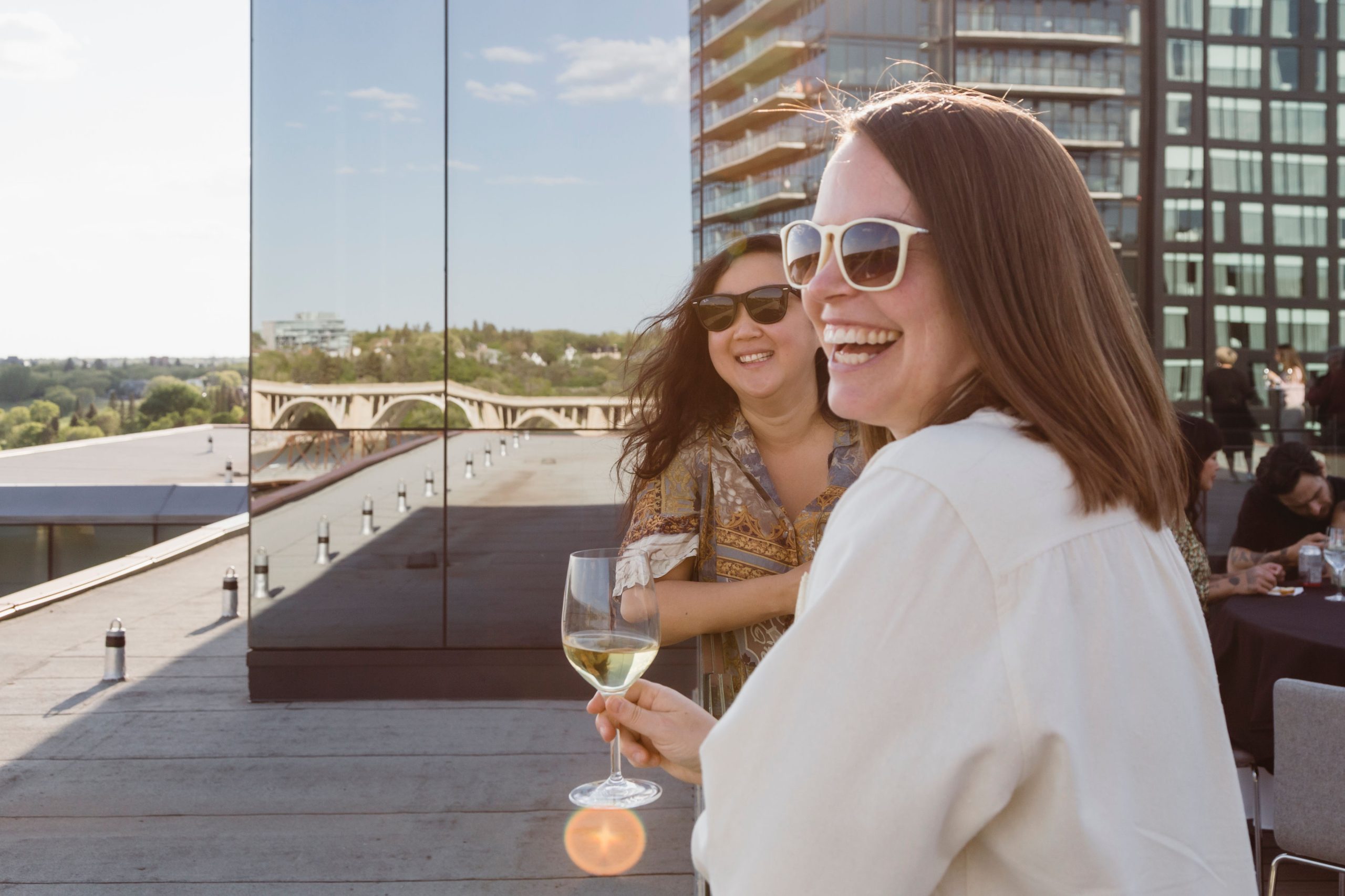 Two people smile and look out at the view from Remai Modern's rooftop. One person holds a wine glass in their hand.