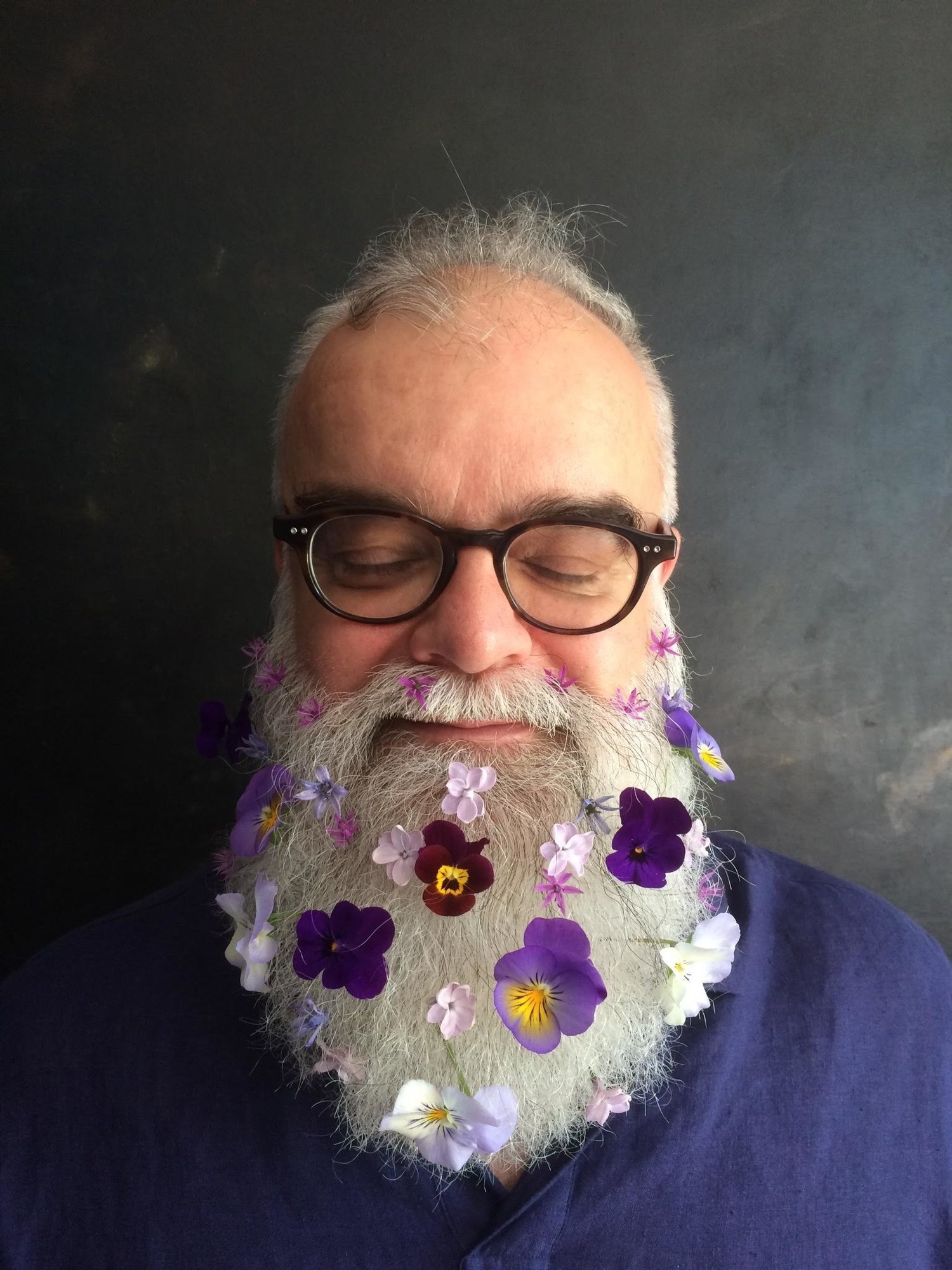 A portrait of composer Rodney Sharman shows him wearing thick-rimmed glasses and sporting a long grey beard with purple flowers in it.