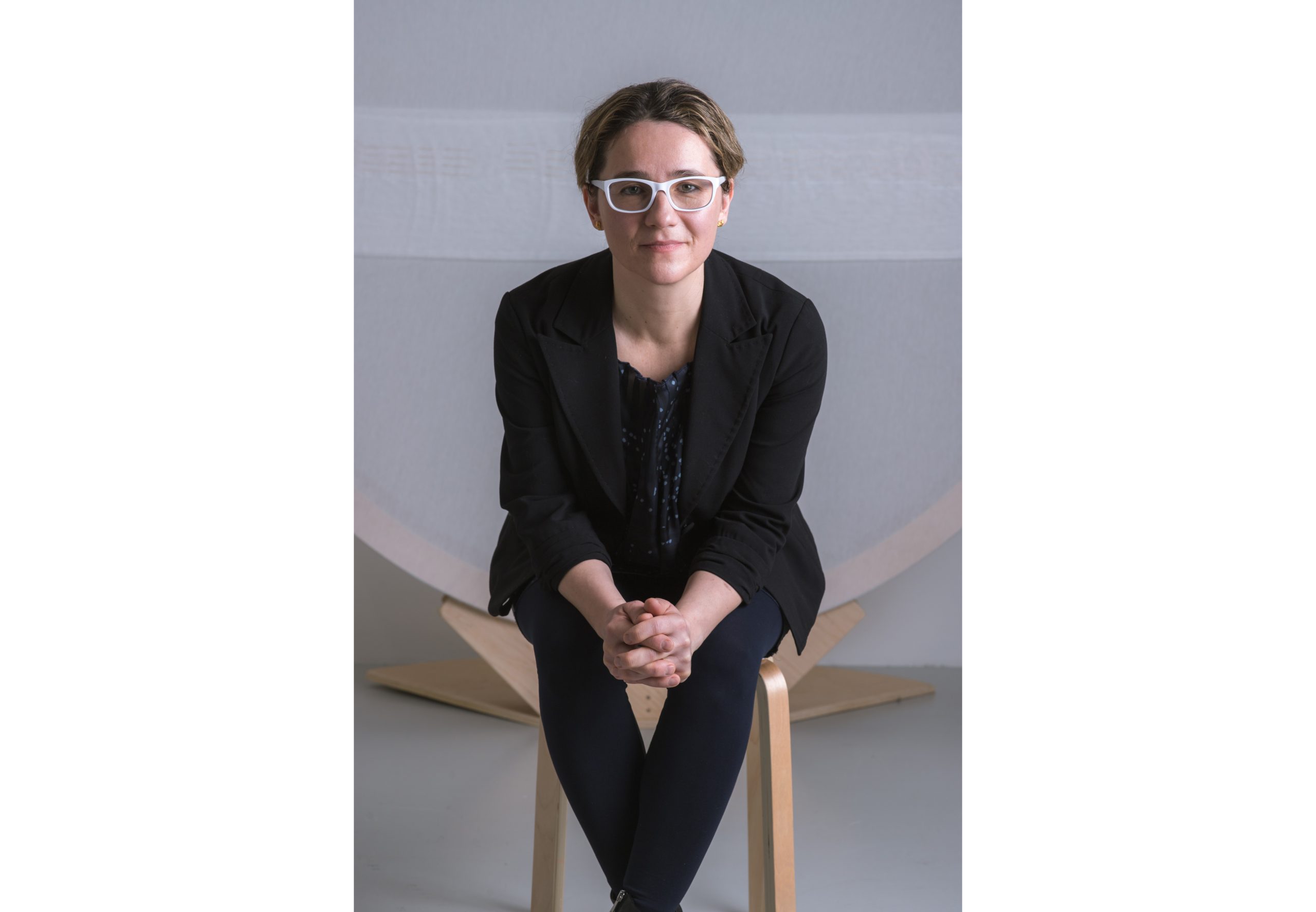 "Portrait of Adelina Vlas, seated and facing the camera, wearing white-framed glasses and a black blazer. Photo by Max Power for D.PE Agency."