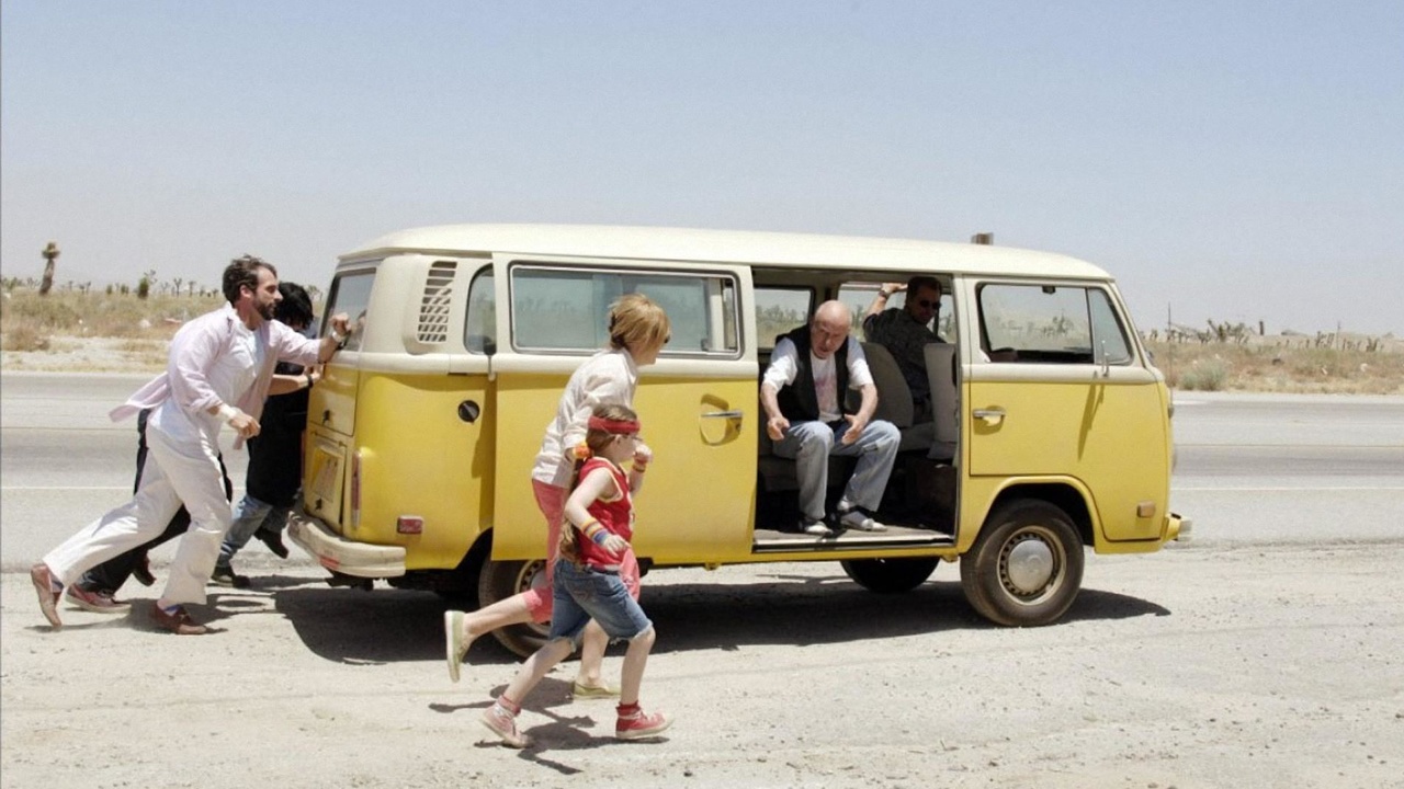 A group of people pushing a yellow Volkswagen van on a dusty road in a desert setting. The van’s sliding door is open, revealing an elderly man sitting inside and another man behind the wheel. A young girl in a red tank top and headband runs alongside the van, while two adults, one wearing a white shirt and the other with blonde hair, push from the rear. The scene captures a sense of teamwork and urgency under the bright sun.