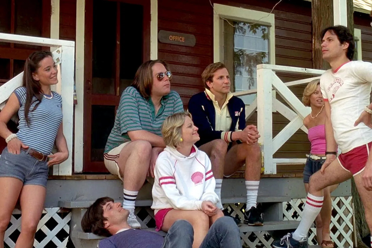 A group of young adults in casual summer clothing is gathered on the steps of a rustic camp building. They appear relaxed and are engaged in conversation. The individuals include a woman in a blue striped shirt and denim shorts, a man in a green striped polo shirt and shorts, a woman in a white hoodie with a red design, a man in a navy jacket with a white shirt, another woman in a pink tank top and denim shorts, and a man in a white T-shirt and red shorts standing with his hands on his hips. The setting is casual and suggests a summer camp environment.