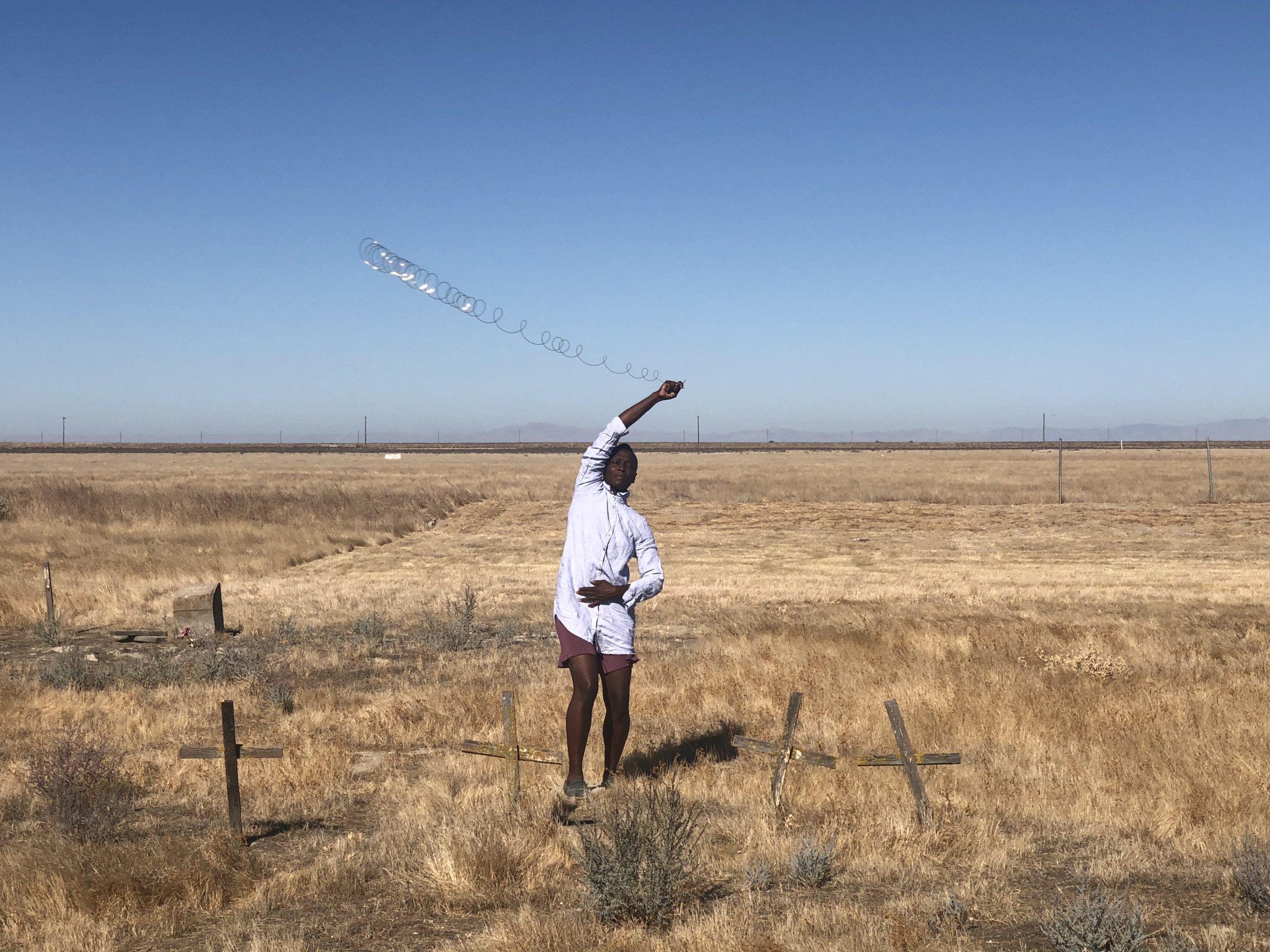 A photo of artist taisha paggett standing in a field covered in dry grass and a few weathered wooden crosses. The artist is wearing a light blue, long-sleeved button-up shirt and brown shorts and is swinging a coiled piece of wire around her head.