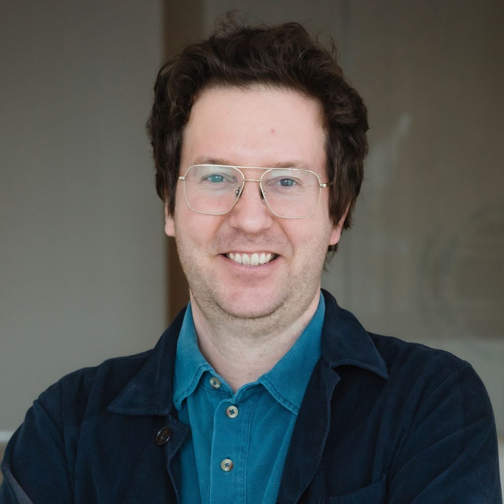 A person with short, wavy dark hair and light-colored glasses smiles at the camera with their arms crossed. They are wearing a teal buttoned shirt under a dark jacket and are standing indoors against a softly blurred background.