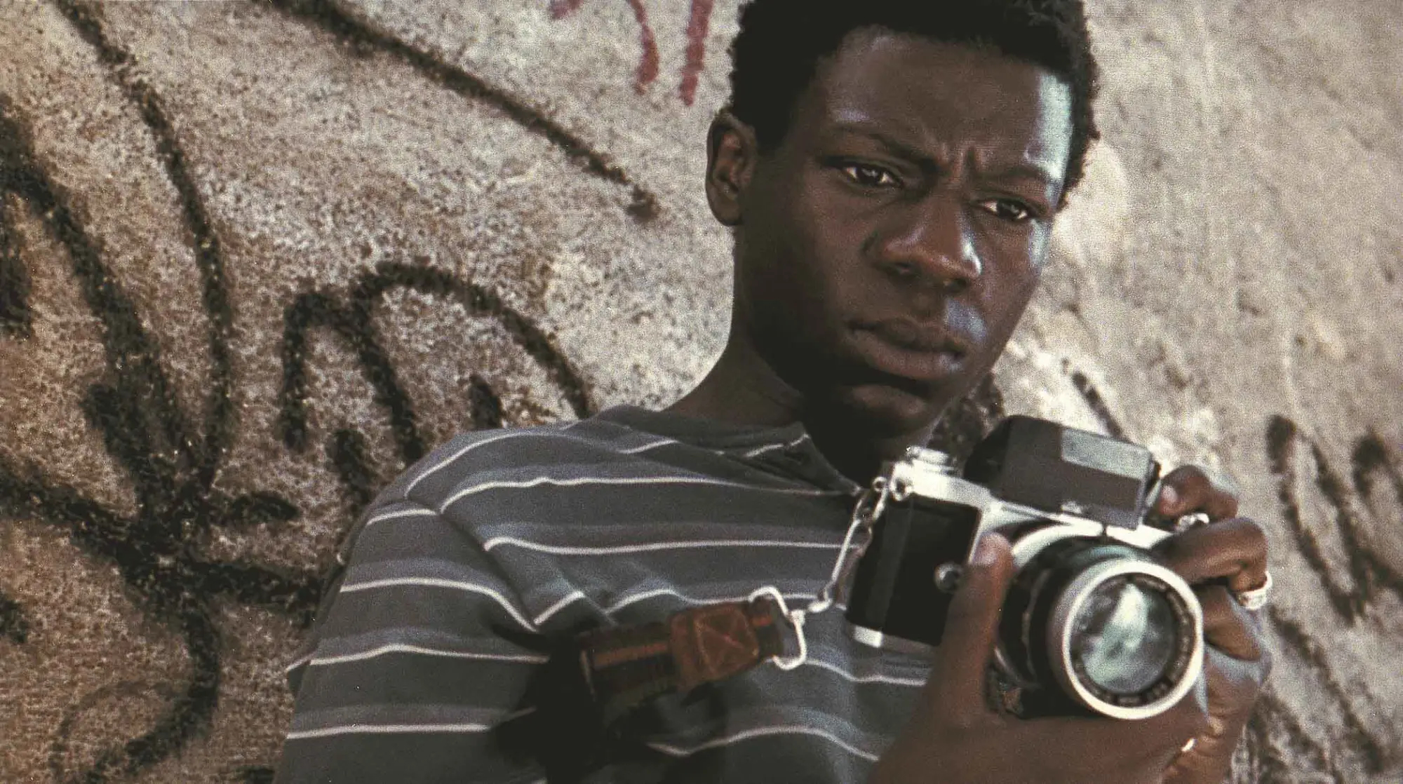 "A young man with a serious expression, wearing a striped shirt, holds and examines an old camera."