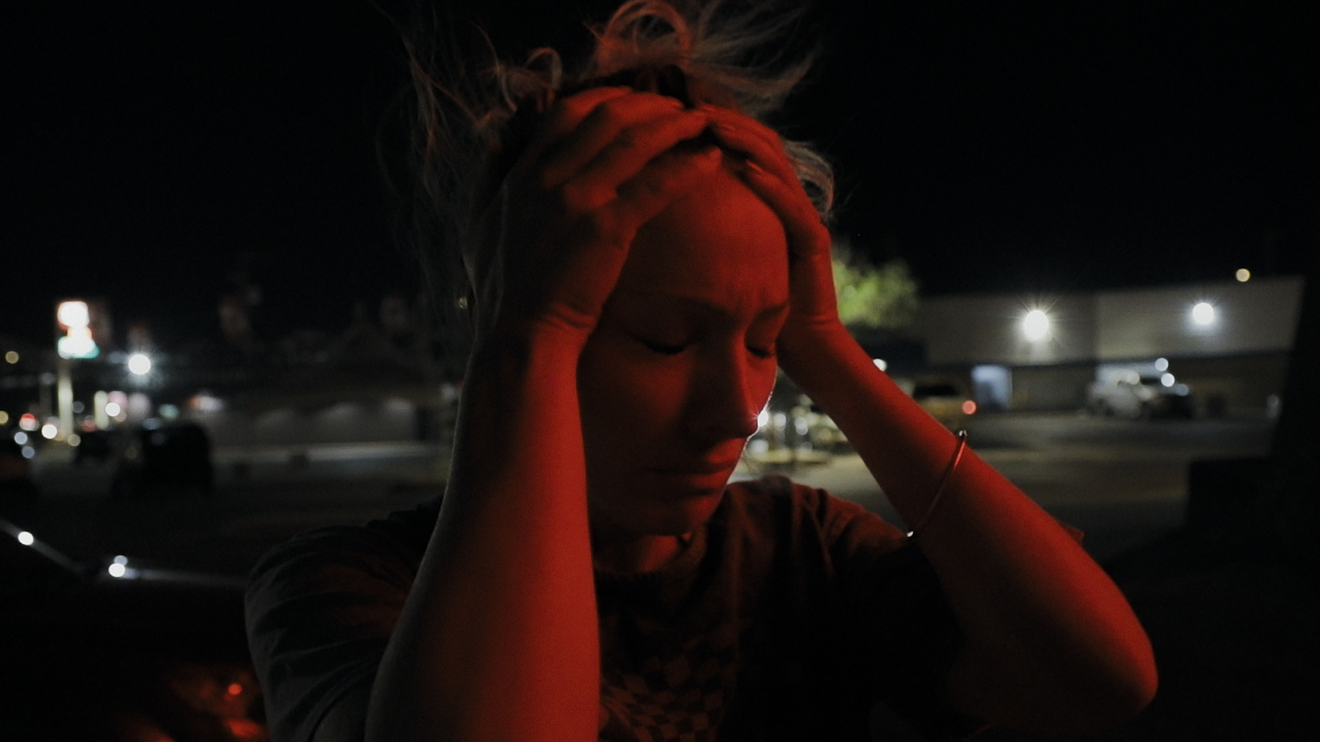 A person holding their head, seemingly in distress, illuminated by a red light in what appears to be a nighttime setting.