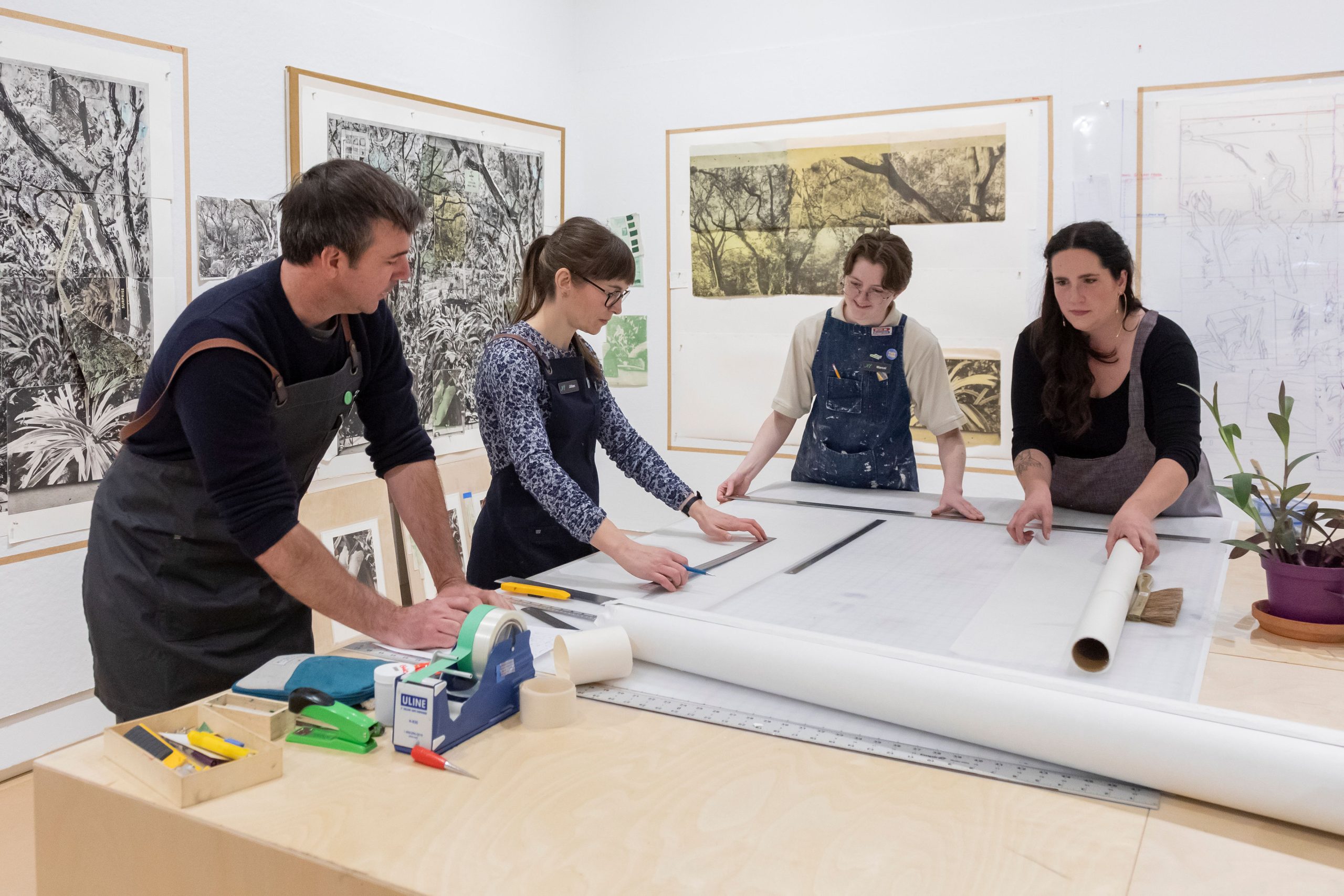 Four individuals collaborating in an art studio, working together on a large piece of artwork. The table is filled with various tools, such as tape, rulers, and cutting instruments, while the walls are adorned with detailed, nature-themed prints. The group appears focused and engaged in their creative process.