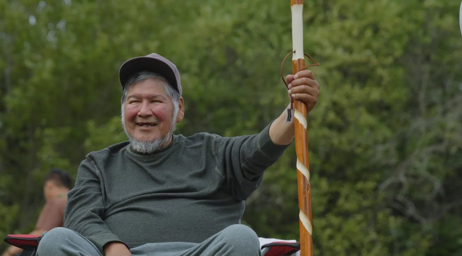 "An older man with a beard and a baseball cap smiles warmly while holding a staff with decorative patterns."