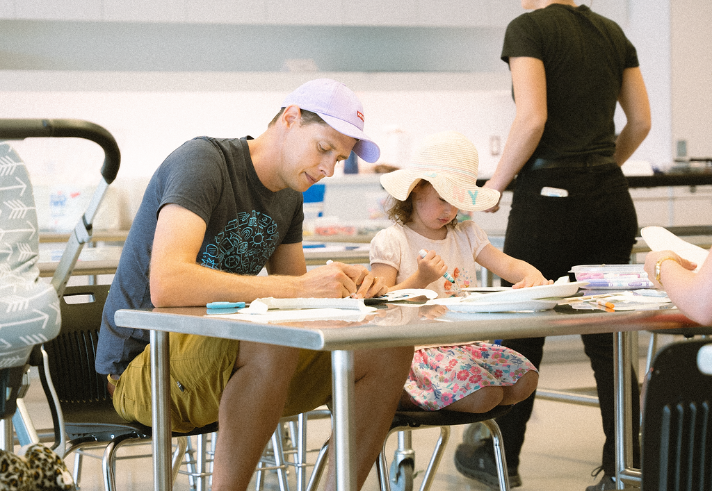 A man and a young child seated at a table, both engaged in an art activity in a classroom or workshop setting.