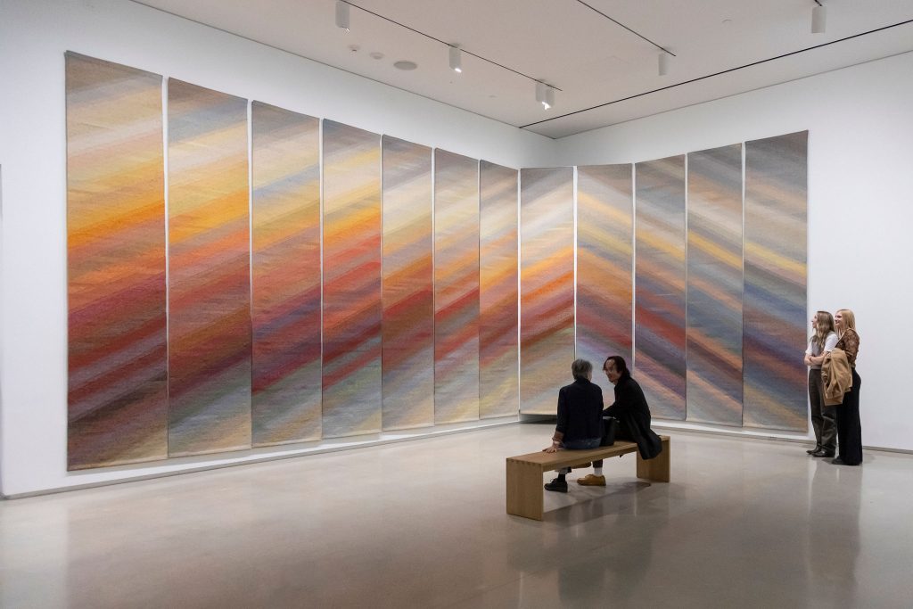 A large, multi-panel textile installation displayed in a gallery, with soft diagonal gradients shifting from warm oranges and reds to cool blues and greys across each panel.