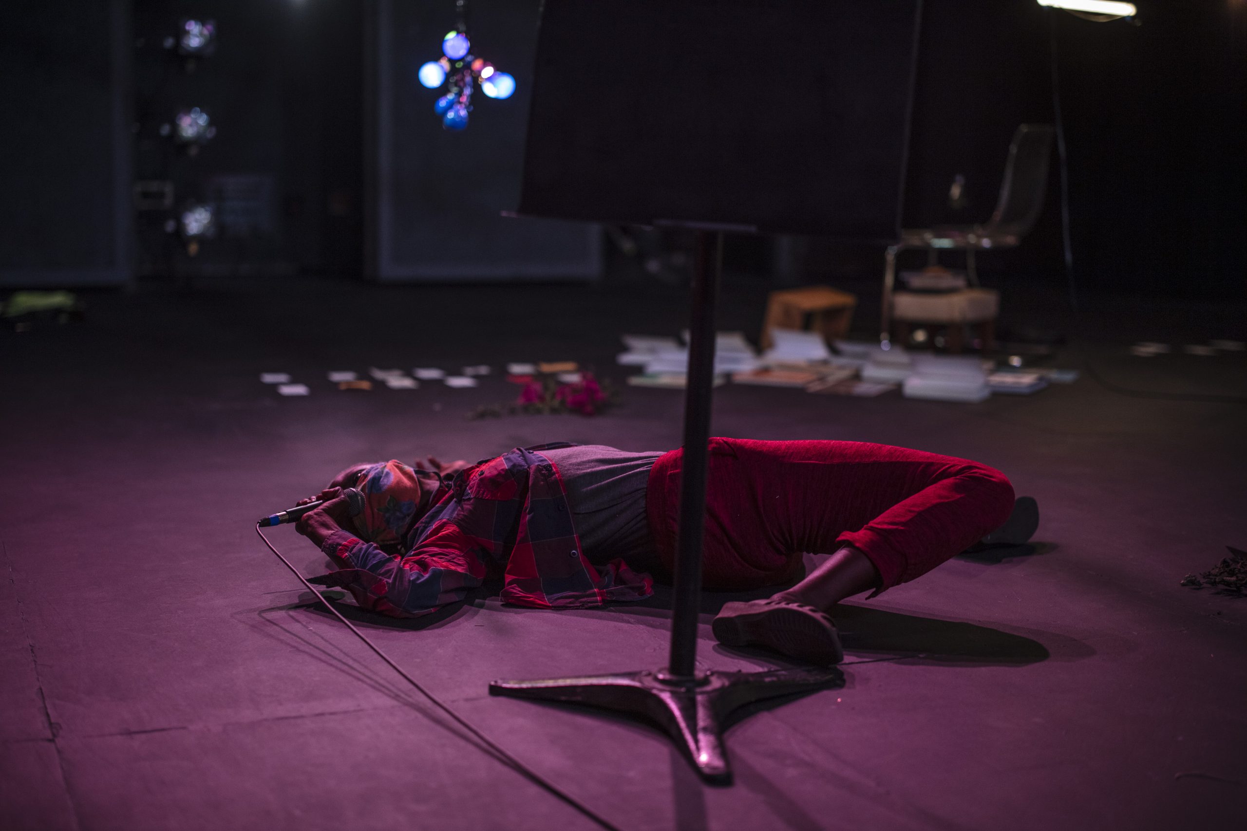 This image shows artist taisha paggett during a performance. She is photographed in a dark gallery space with low lighting and is lying on the floor holding a microphone.