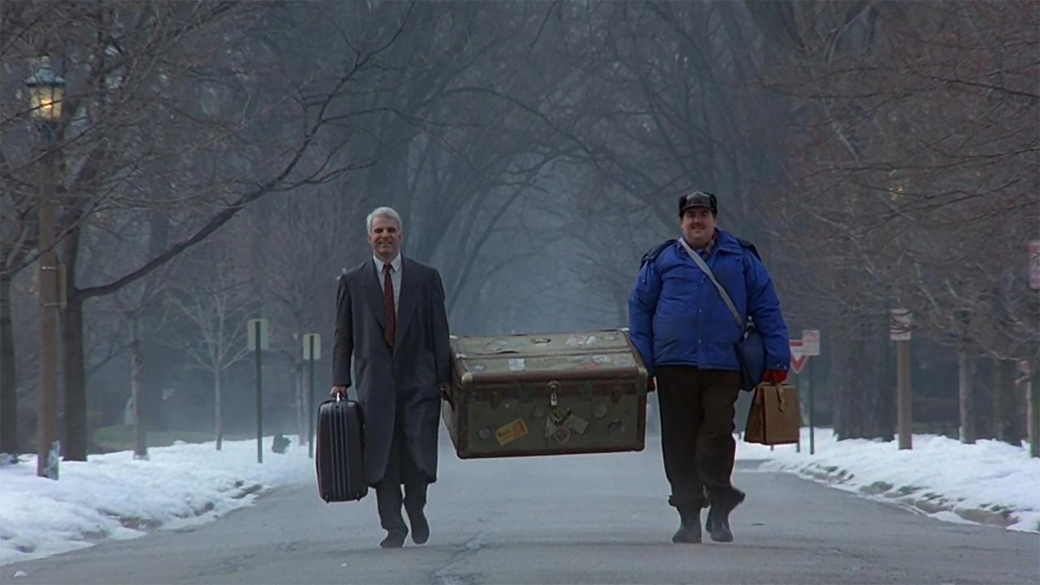 "Two men walking down a snowy street carrying large luggage together."