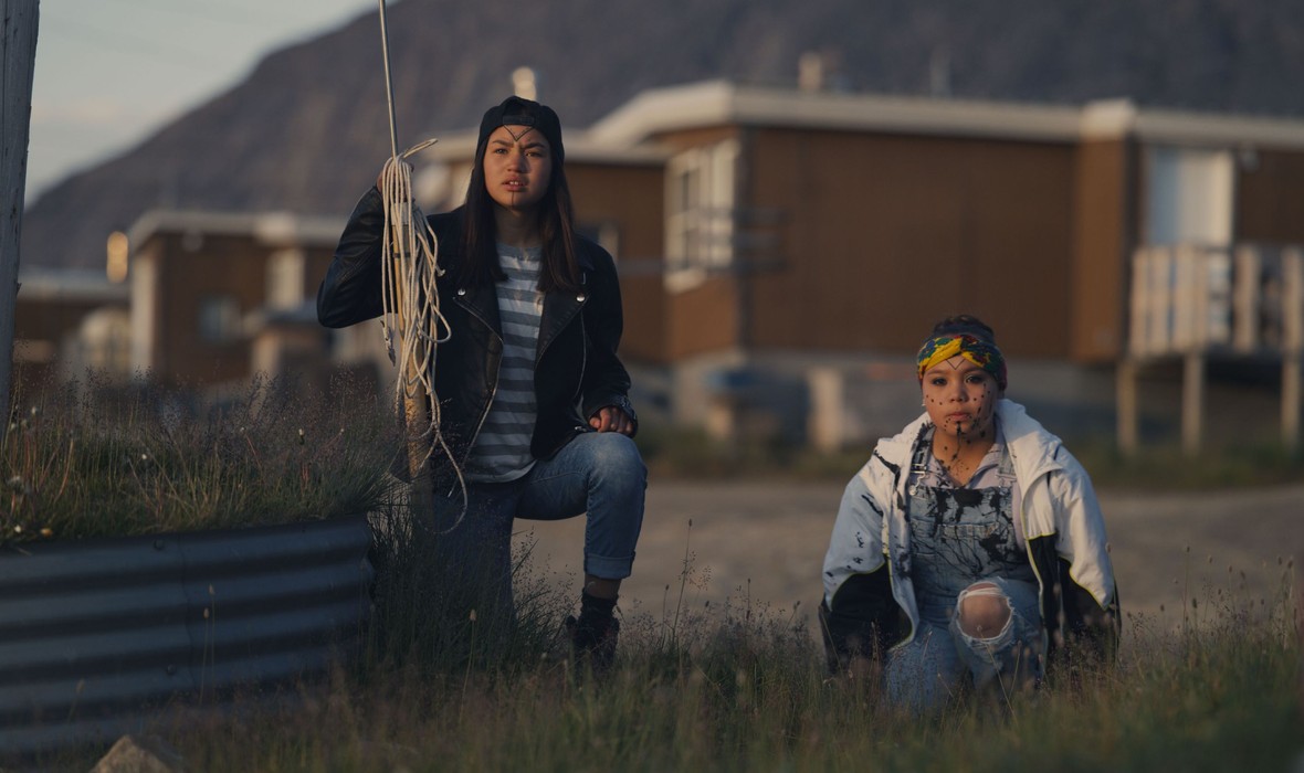 "Two young women crouch outside in a grassy area, one holding a rope, with houses and mountains in the background."