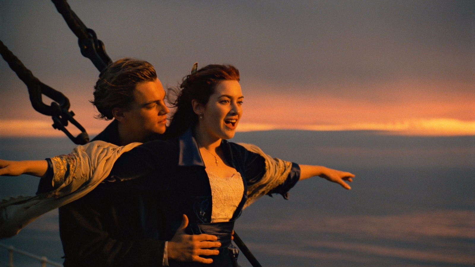 "A loving couple stand on a sailing boat in the middle of the ocean facing the sunrise with his hands on her waist".