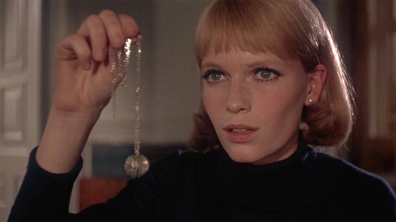 "A close-up of a woman with short blonde hair and a concerned expression, holding up a necklace with a pendant."