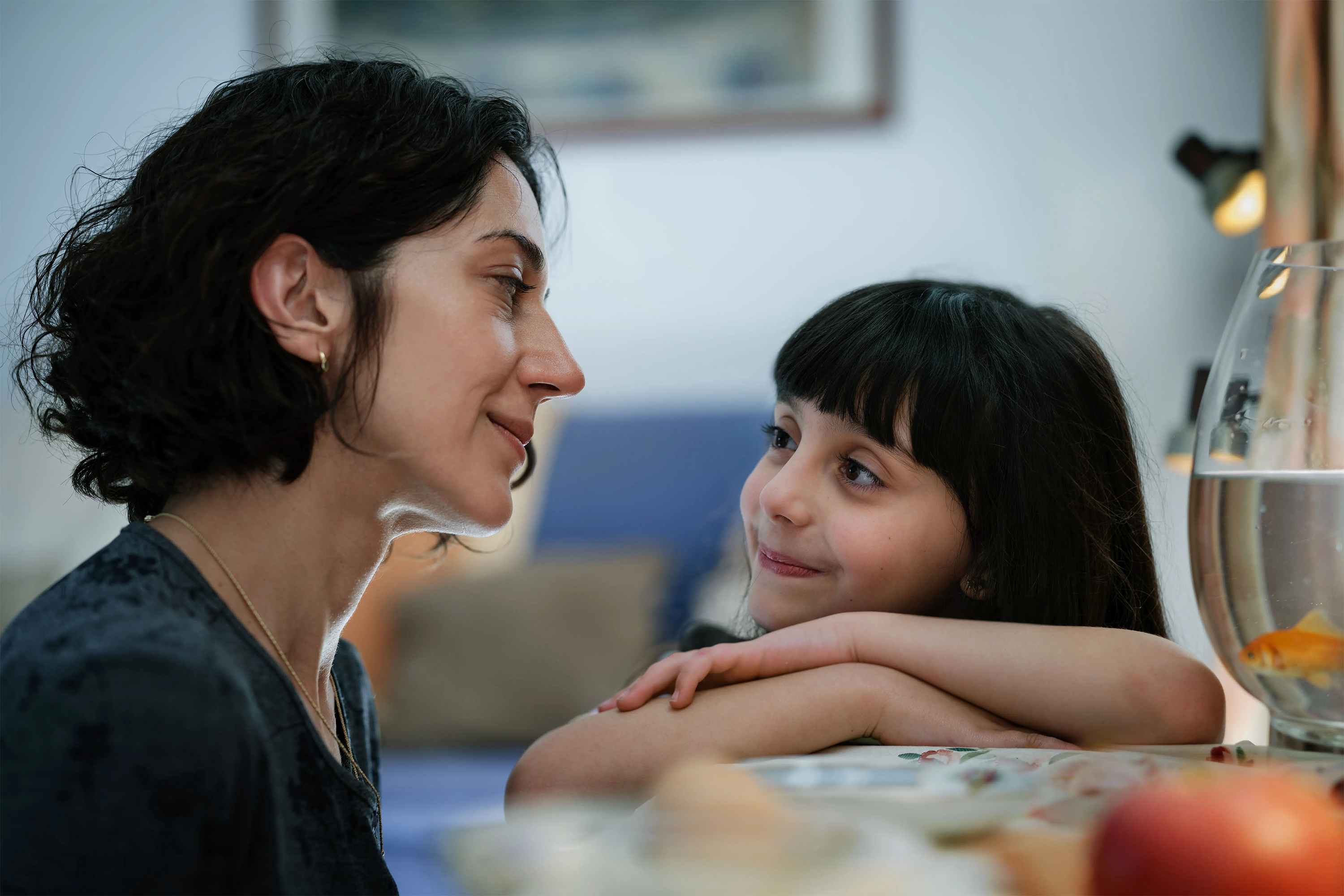 "Woman and young girl smiling at each other across a table with a goldfish bowl nearby."