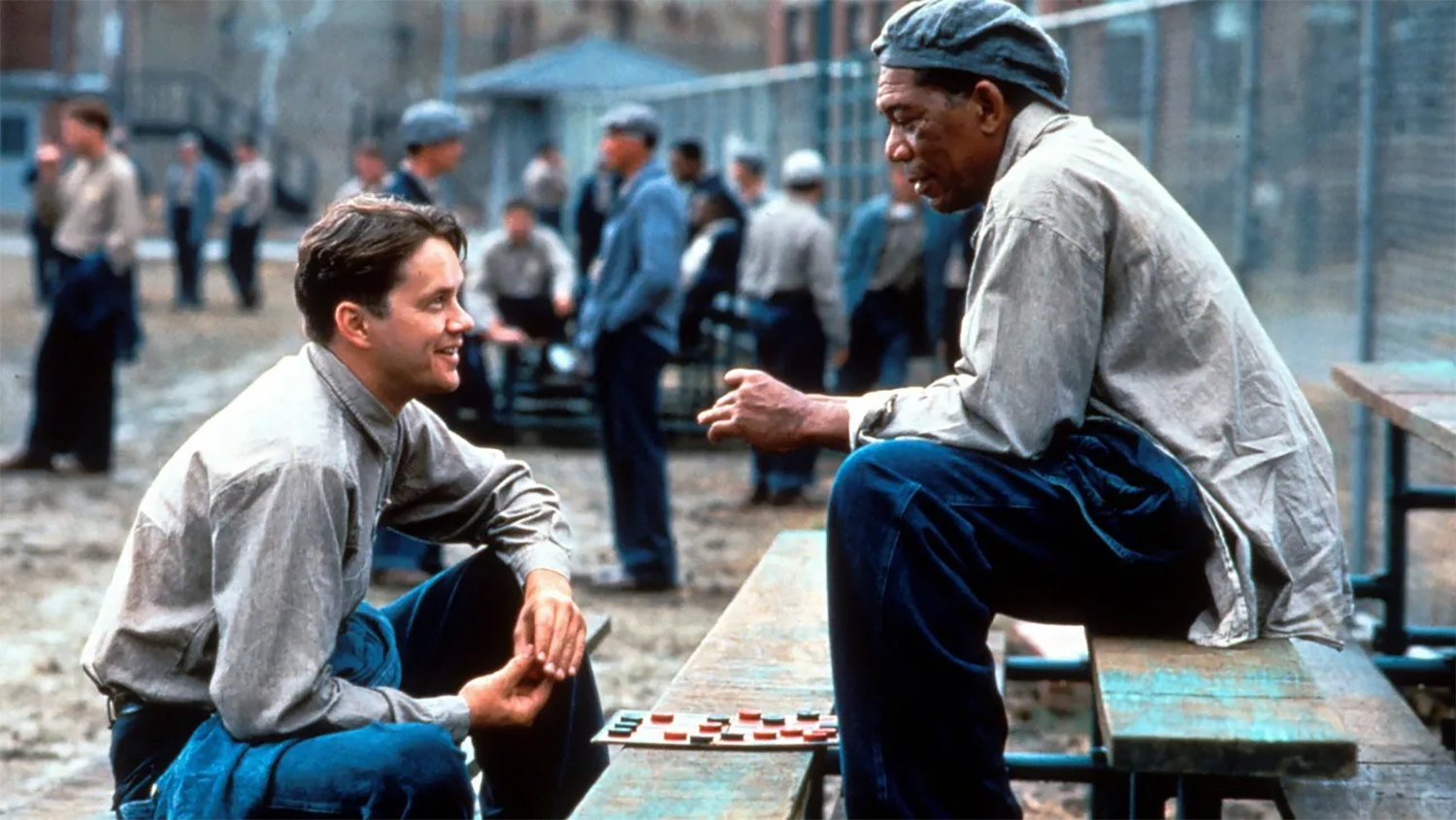 Two men in prison uniforms sit outside on a bench, talking and playing checkers.