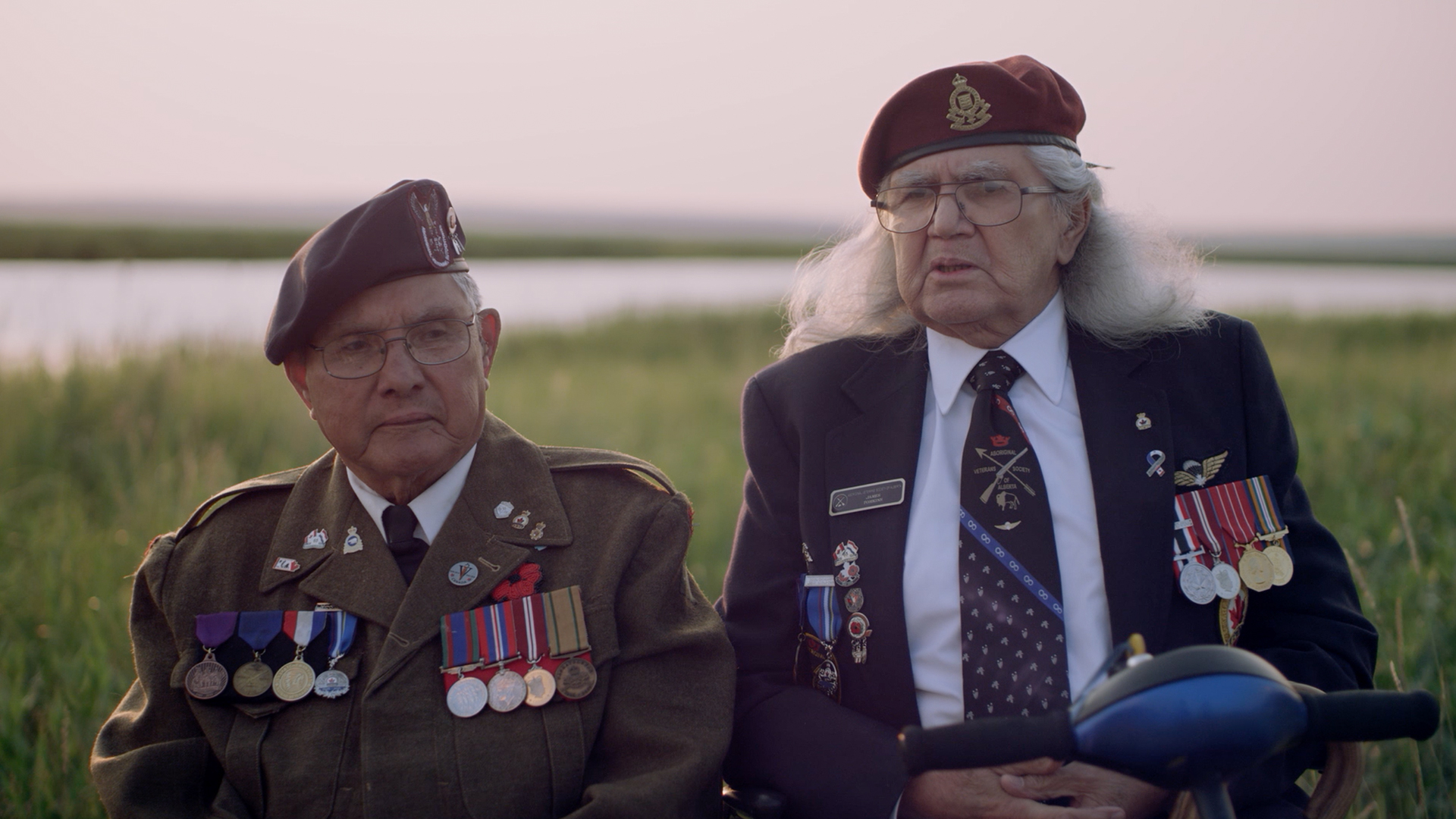 This image shows two elderly veterans sitting outdoors, possibly near a lake or river, as the backdrop includes grass and water under a soft, warm light. Both individuals are wearing military uniforms and berets adorned with insignias, along with multiple medals pinned to their jackets, suggesting they are decorated war veterans. One veteran, on the left, is wearing a traditional military uniform with several medals, while the other, on the right, has long gray hair, glasses, and is dressed in a blazer, also showcasing medals and military patches on his chest. Their attire and expressions convey a sense of honor and respect for their service.