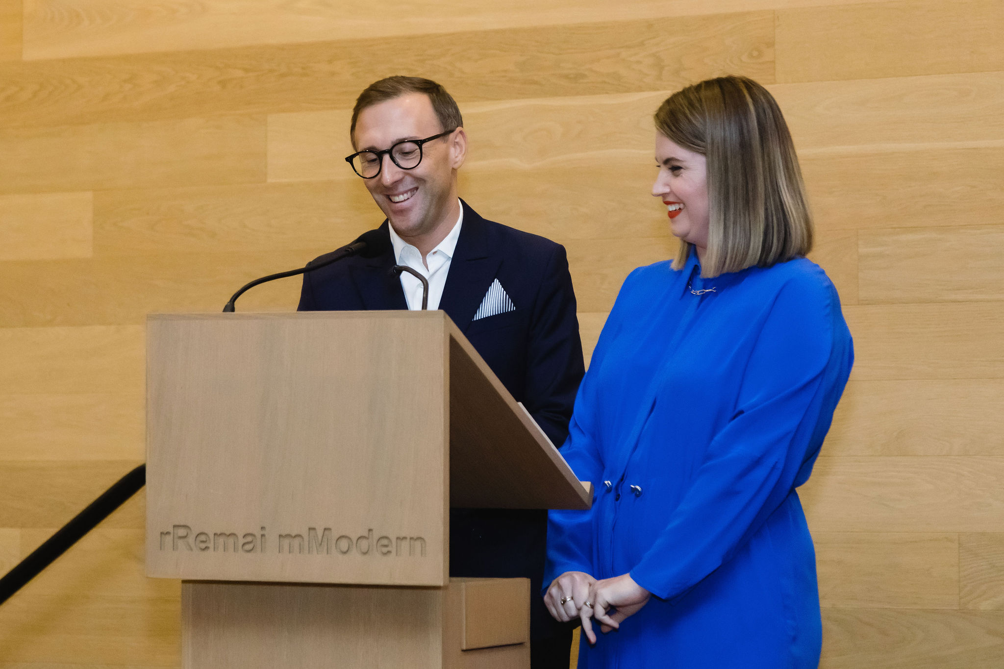 A man and a woman stand at a wooden podium with the Remai Modern logo engraved on it. The man, wearing glasses and a dark suit with a white shirt and pocket square, smiles while speaking into the microphone. The woman, dressed in a bright blue outfit, stands beside him, smiling warmly.