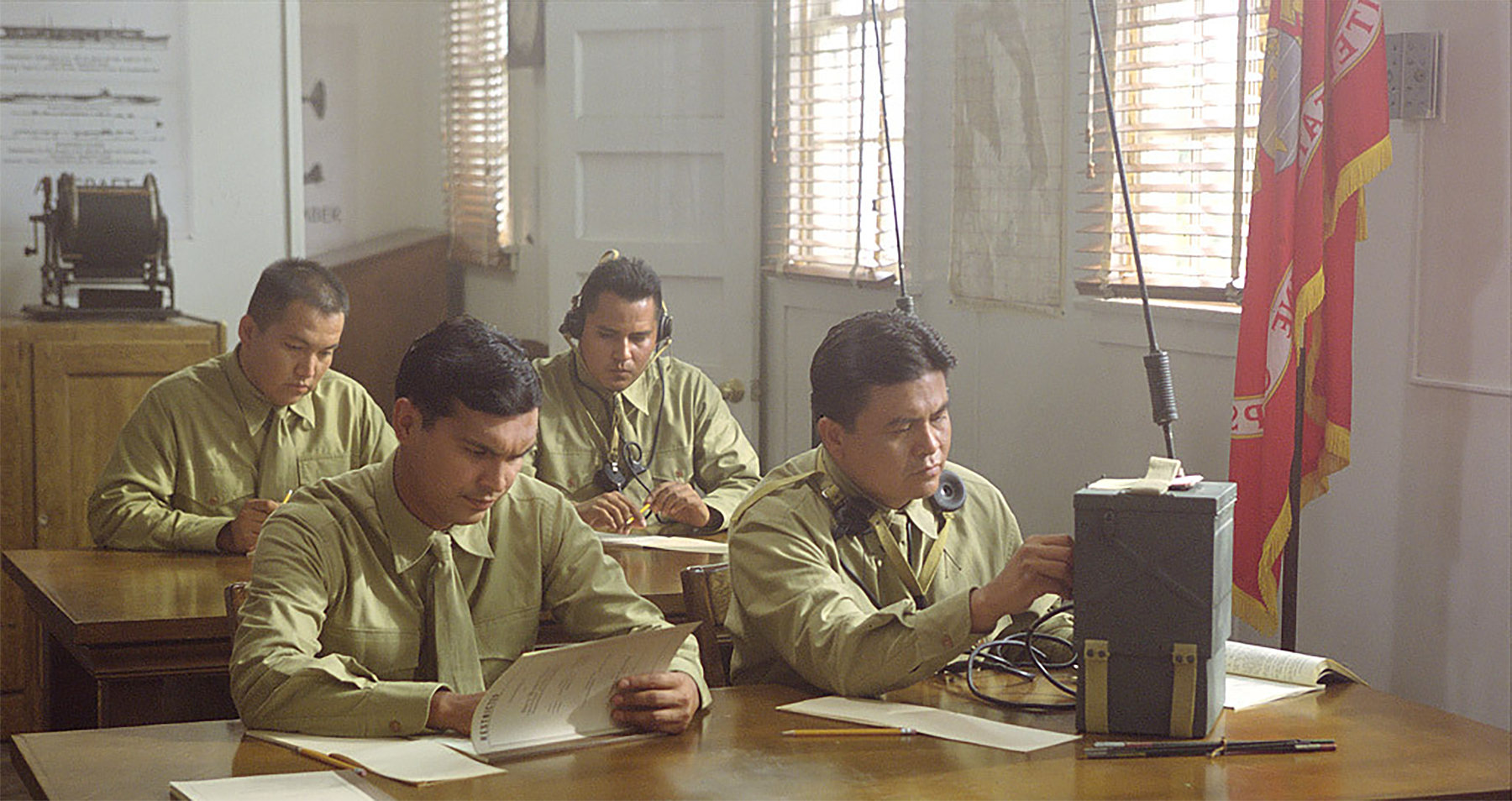 The image shows a group of individuals in military attire working in a classroom-like setting. They are seated at tables, using radios and wearing headphones, possibly engaged in communication tasks. The individuals appear to be focused on writing or reading documents. The setting suggests a historical context, likely related to military communication operations, with the presence of military flags and equipment such as radios.