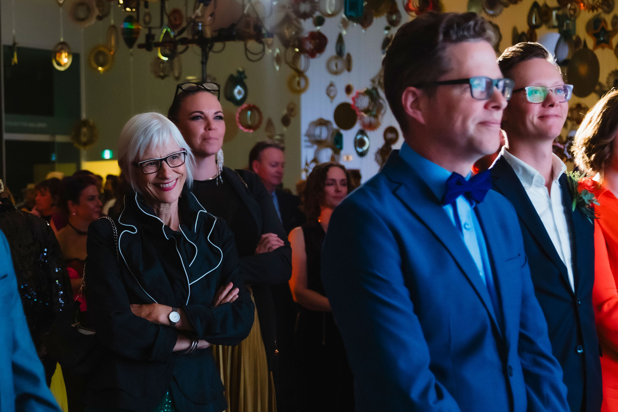 A group of people in a dimly lit room smile and they listen to a speaker not visible in the photo. An artwork made with cascading, colourful shapes can be seen in the background.