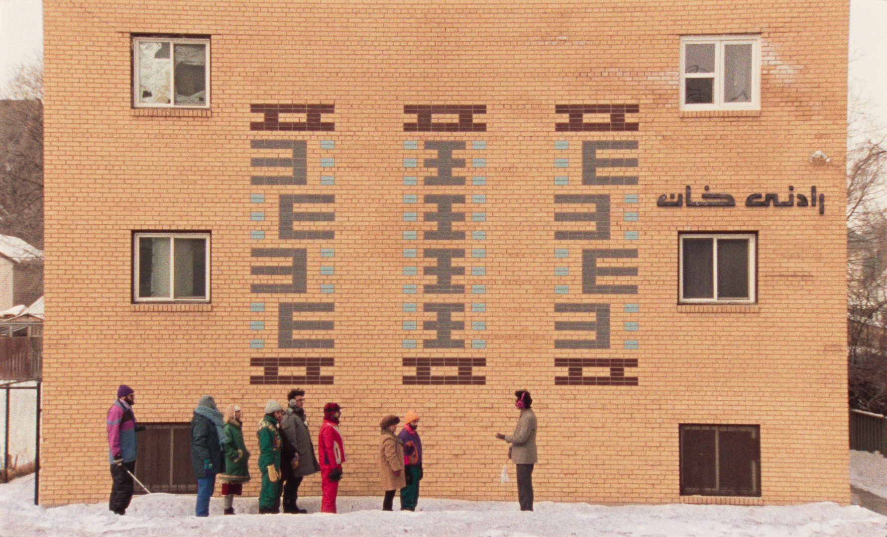 A group of people dressed in colorful winter clothing stand in a line on a snowy ground in front of a yellow brick building. The building features symmetrical patterns made of blue and brown tiles, with some windows and text in a foreign script on the wall. A man at the end of the line appears to be speaking to or addressing the group, holding a white plastic bag in one hand. The scene conveys a quiet, structured moment in a cold, urban setting.