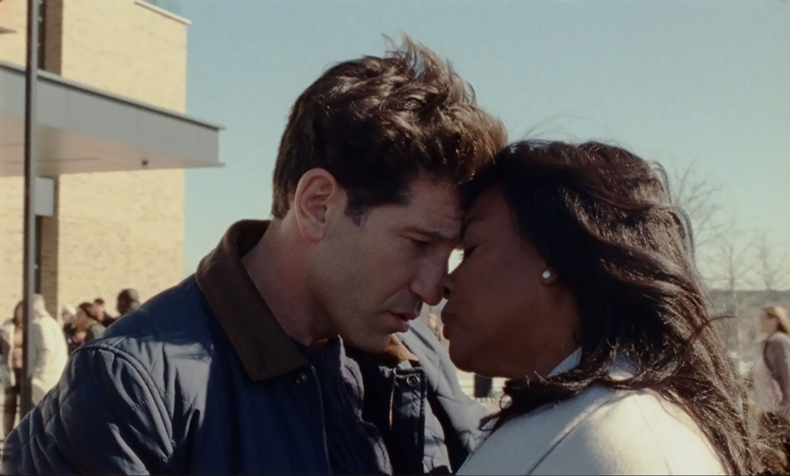 A man and a woman share an intimate moment outdoors, their foreheads gently pressed together as they close their eyes. The man is wearing a blue jacket, while the woman, with pearl earrings and flowing hair, is dressed in a light coat. In the background, a sunny day reveals a building and several blurred figures, suggesting a public setting. The scene conveys a sense of emotional connection and tenderness.