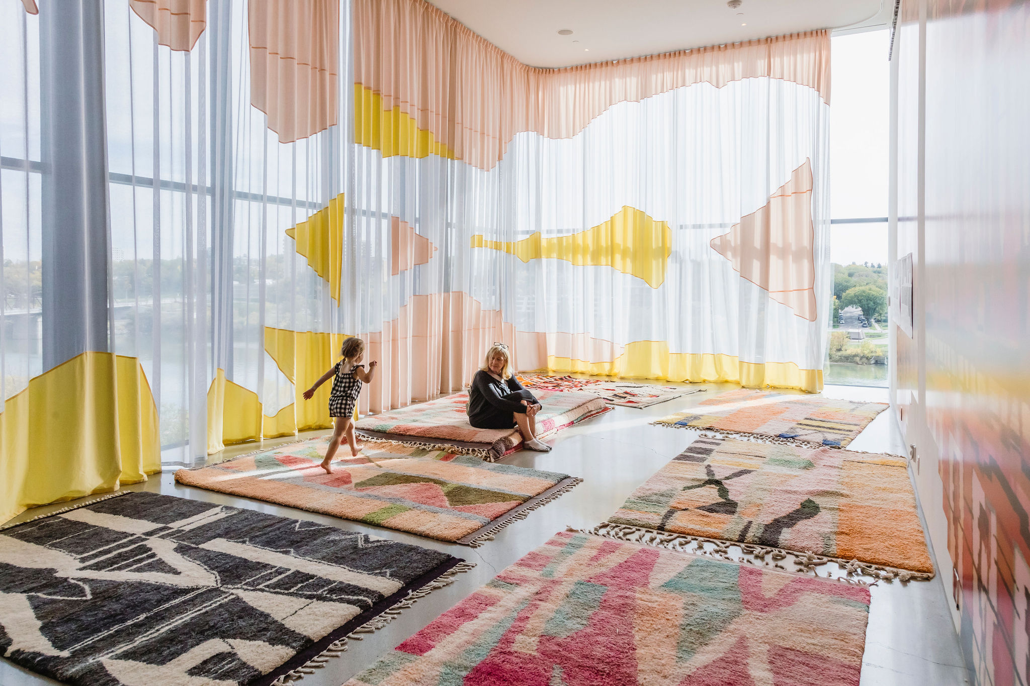 A spacious room filled with colourful rugs and modern artistic curtains. The rugs have various abstract patterns and colors, adding warmth and vibrancy to the space. A woman sits on one of the rugs, and a child is walking or playing nearby, adding a sense of liveliness to the scene. The curtains feature large yellow, pink, and white shapes, and natural light streams through the large windows, creating a soft and inviting atmosphere.