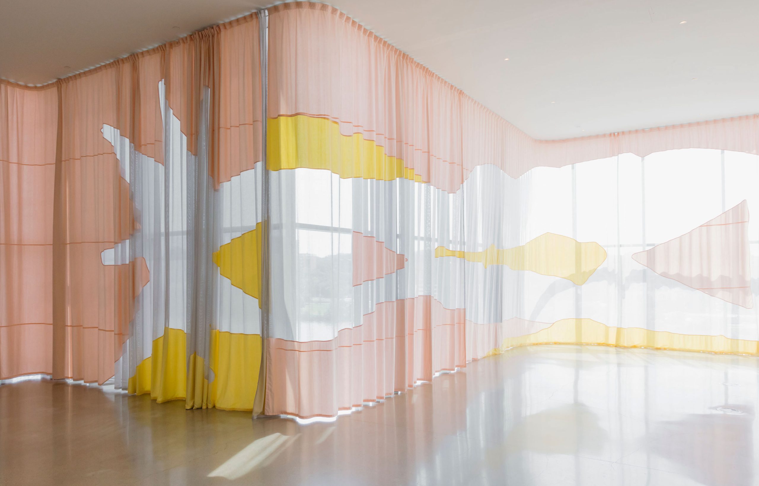 An elegant art installation featuring sheer, floor-to-ceiling textile curtains in soft peach, white, and yellow tones. The translucent panels overlap to create abstract, organic shapes, allowing natural light to filter through. The fabric is arranged in gentle curves and folds, enhancing the interplay between light and shadow. The minimalist space, with polished floors and a backdrop of large windows, creates a serene and modern atmosphere, highlighting the delicate and flowing nature of the artwork.