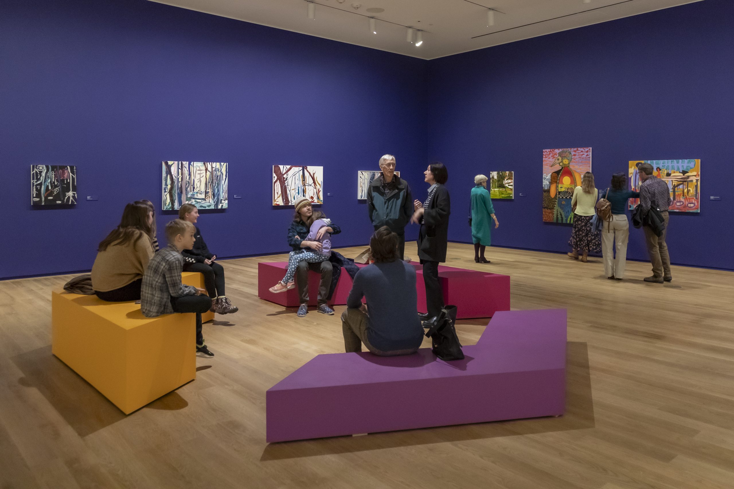 A group of people sit around a tour guide in a colourful gallery exhibition space.