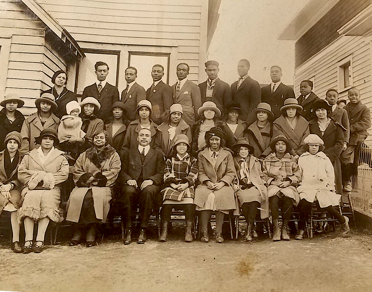 A vintage black-and-white group photograph of well-dressed men, women, and children posed in front of a wooden building. The individuals wear formal attire typical of the early 20th century, including suits, dresses, and hats. The arrangement is orderly, with seated individuals in the front row, followed by standing adults and children in the back rows. The photo captures a sense of community and historical significance.