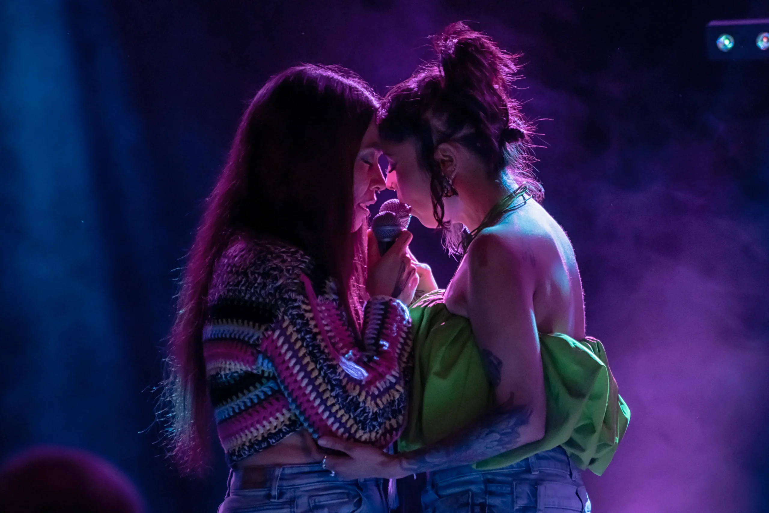 Two performers stand close together on stage under purple and blue lighting, foreheads touching as they share a microphone in an intimate moment during a live performance. One wears a colorful crochet top, the other a green strapless outfit.