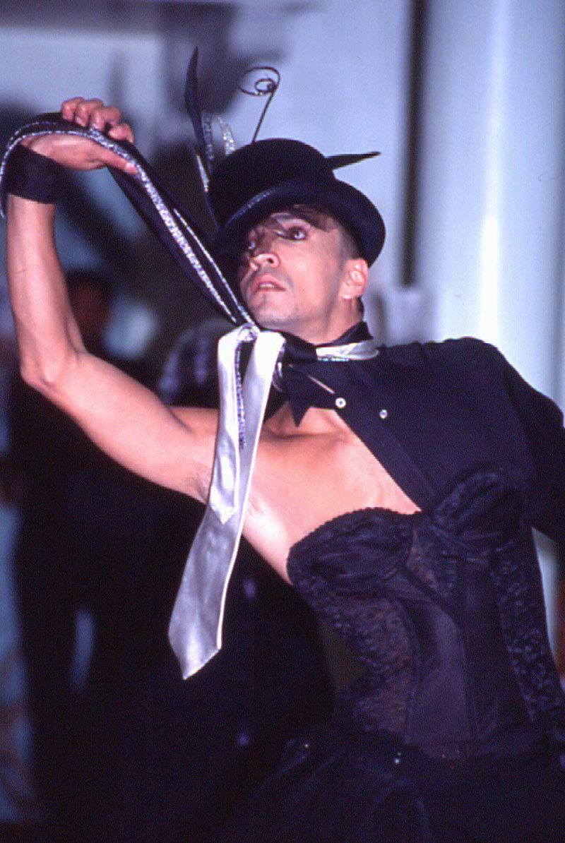 A performer strikes a dramatic pose during a ballroom event, wearing a black corset, a structured top hat with decorative elements, and a silver tie loosely wrapped around their neck. Their arm is extended elegantly, holding the end of the tie, and their makeup is bold and expressive, emphasizing the intensity of their gaze. The background is softly blurred, drawing focus to the powerful stance and theatrical attire.