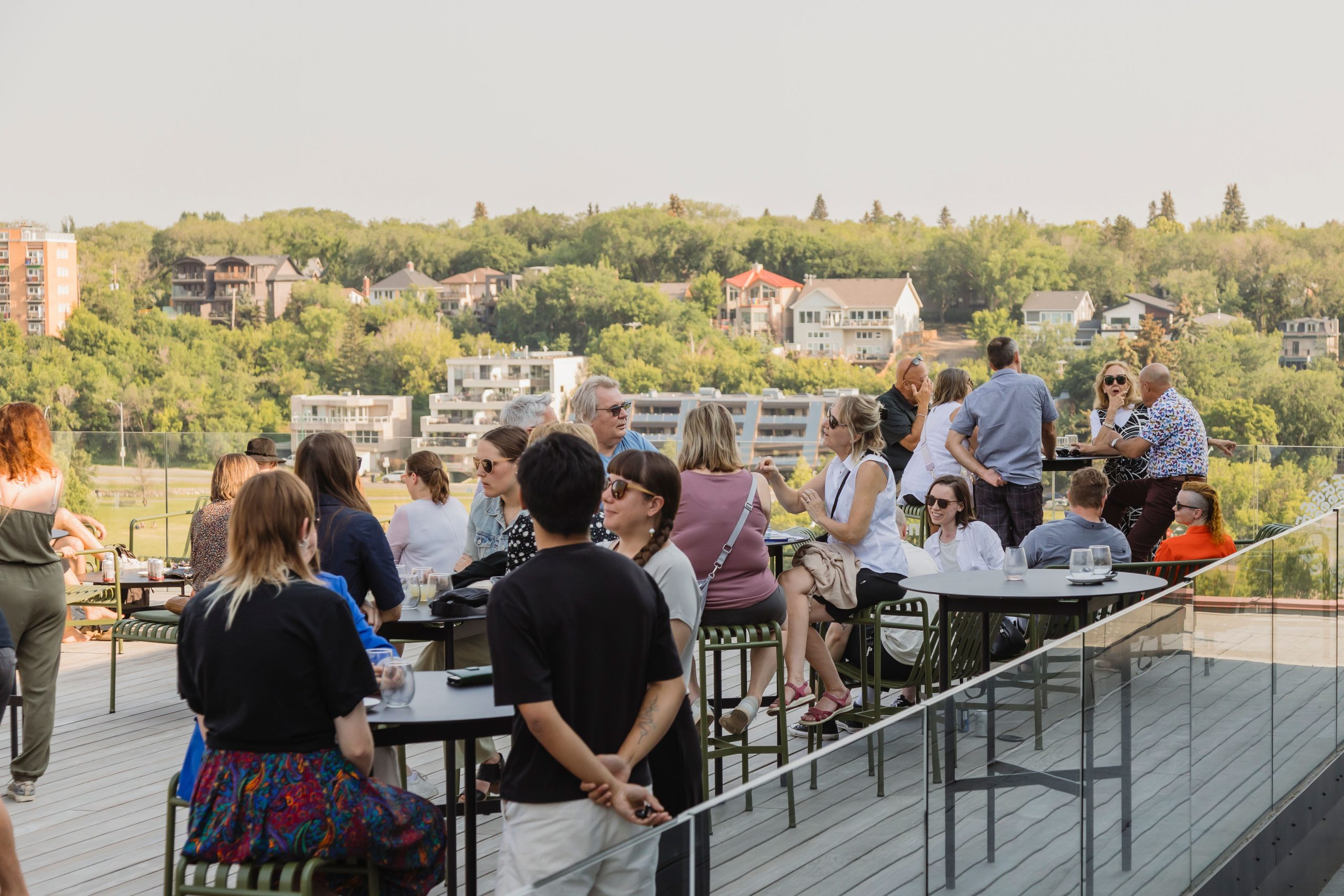 People enjoying drinks and conversation on a rooftop patio during a sunny afternoon, with a scenic view of trees and residential buildings in the background.