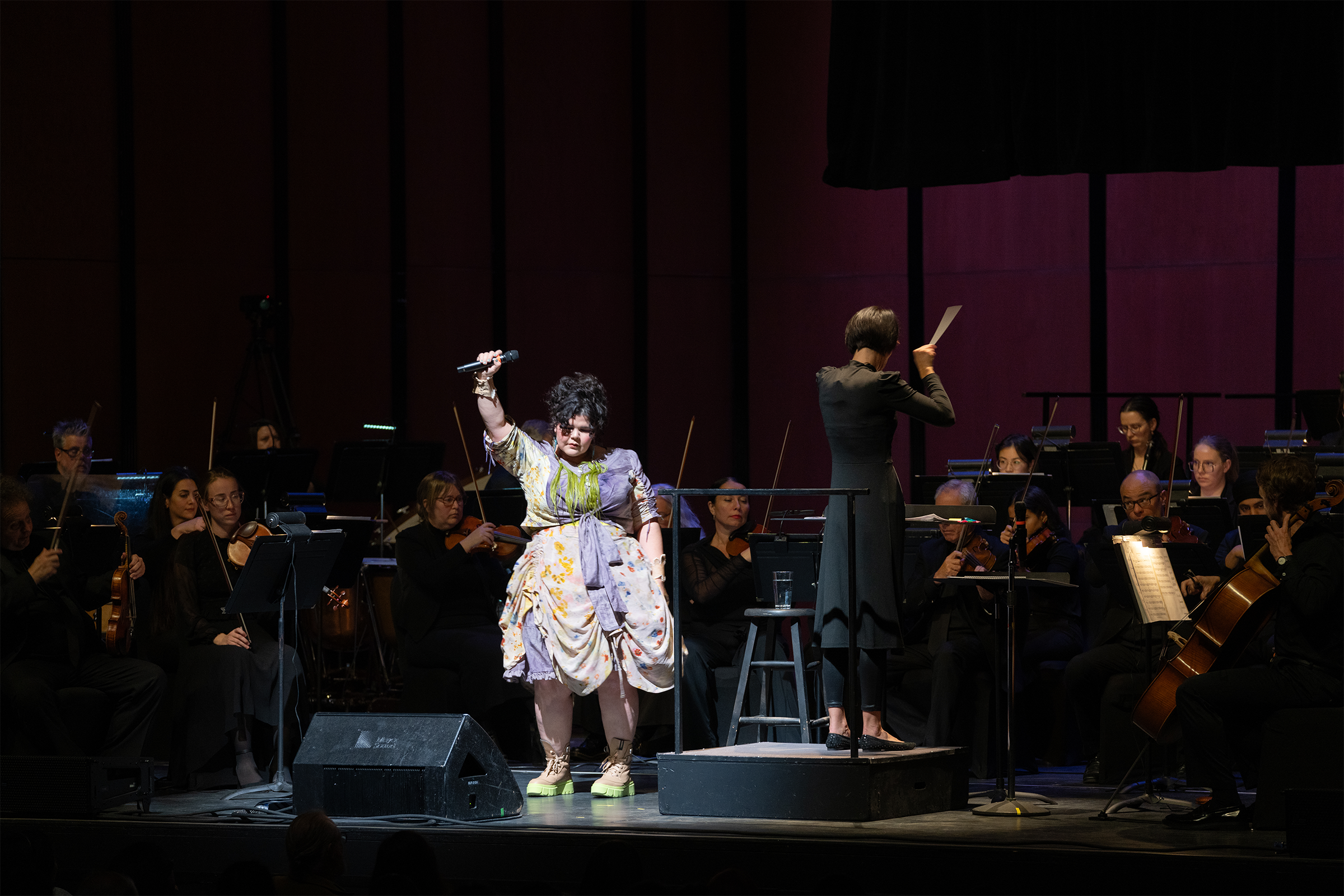 A dramatic performance on stage features a vocalist in a colourful, layered dress and bright green platform shoes holding a microphone high in one hand. She stands in front of an orchestra with string players seated behind her. A conductor in a long black dress stands on a podium, facing the orchestra with a score in hand. The stage is warmly lit against a dark red and black backdrop.