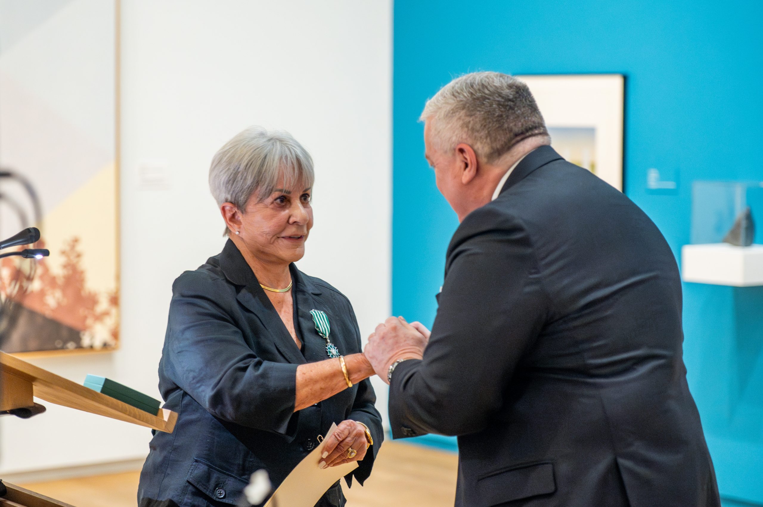 A woman with short gray hair, wearing a dark suit and a medal pinned to her lapel, receives a formal handshake from a man in a suit during an award ceremony at an art gallery.