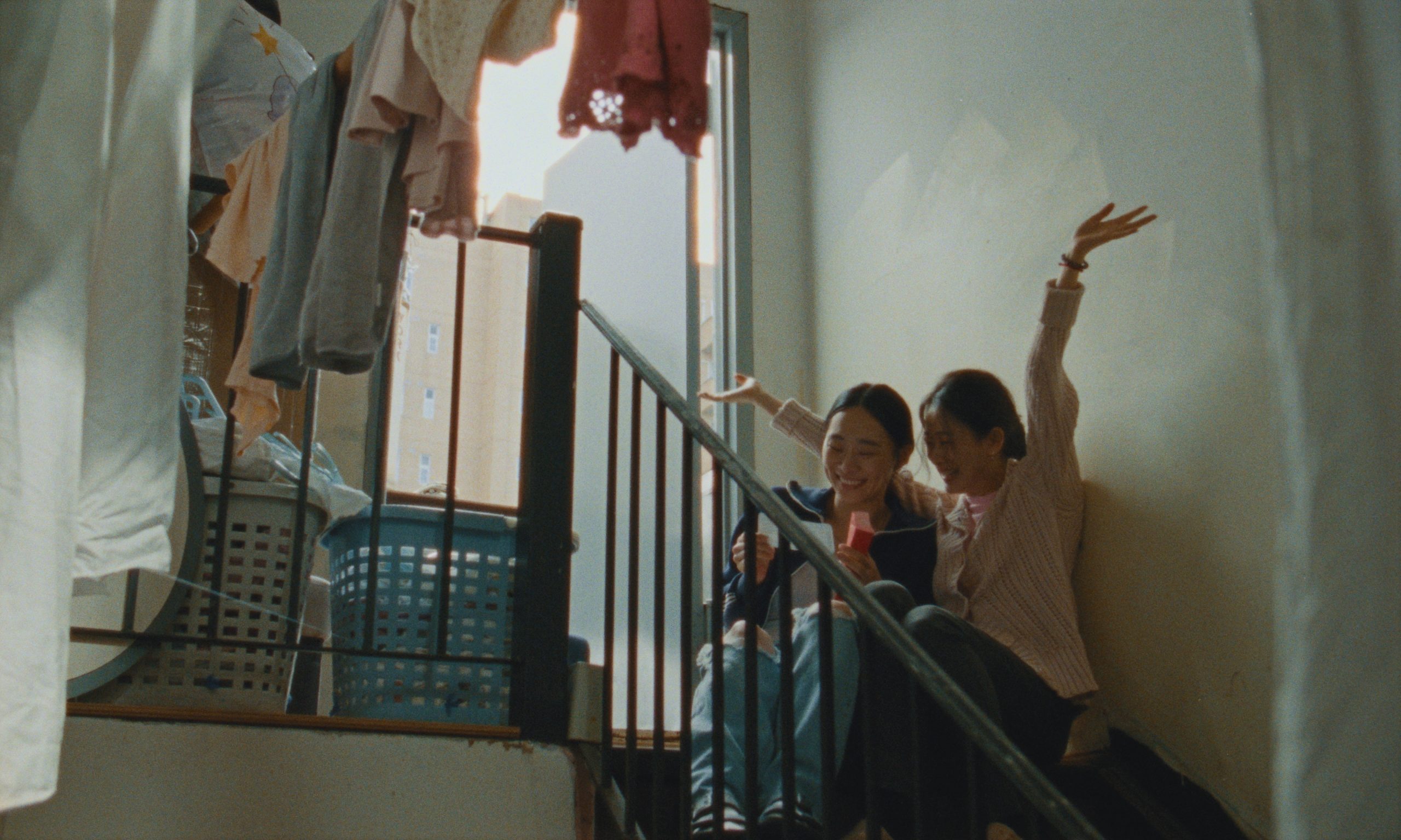 Two young women sit side by side on an indoor stair landing, smiling and laughing together. One holds up a piece of paper while the other raises her arm in excitement. Around them, laundry baskets and clothes hang drying, with warm sunlight streaming through a window.