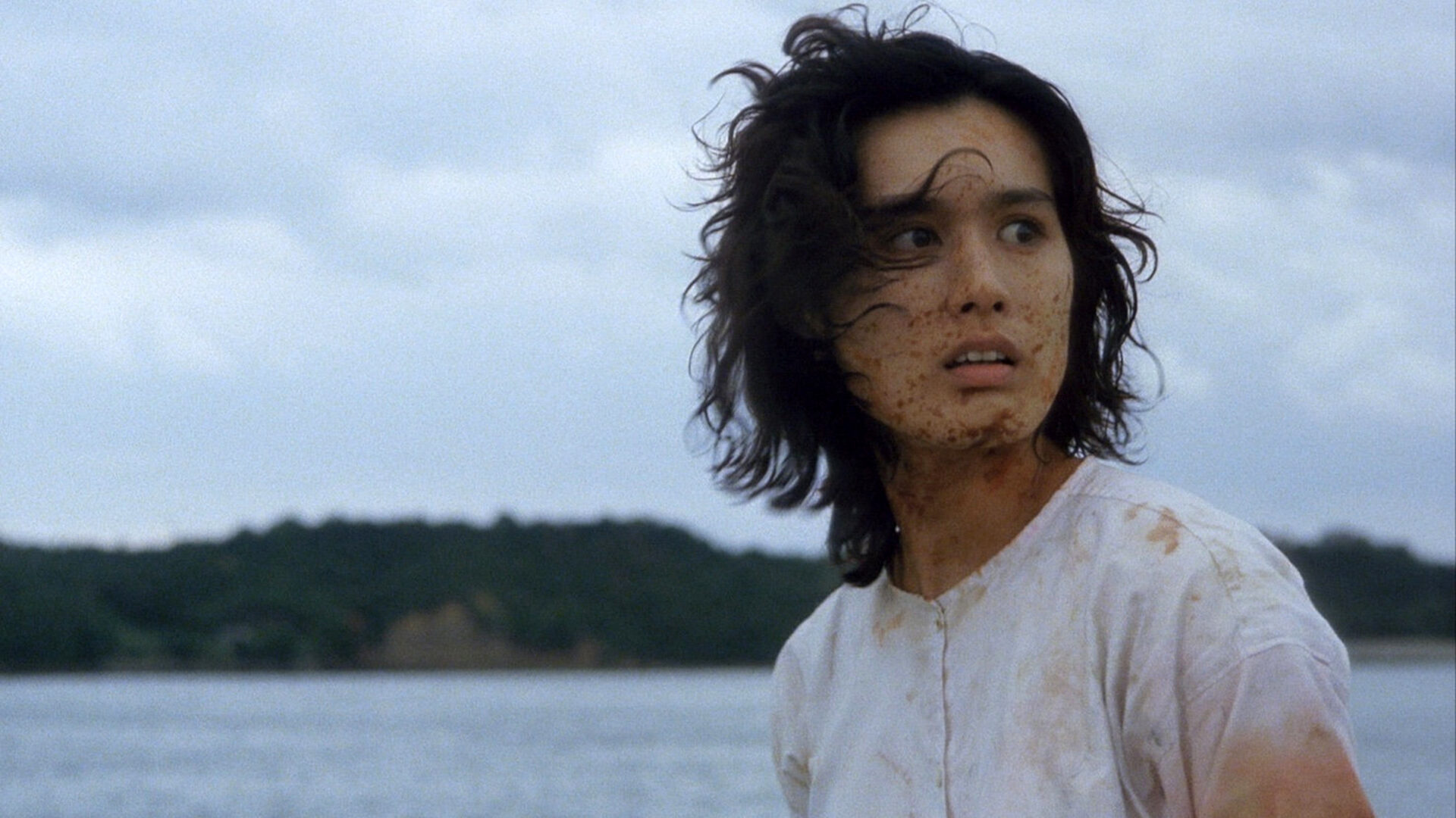 A woman with short, disheveled hair and a fearful expression stands near the water’s edge. Her face and white shirt are splattered with blood. In the background, a wooded shoreline is visible under a cloudy sky.