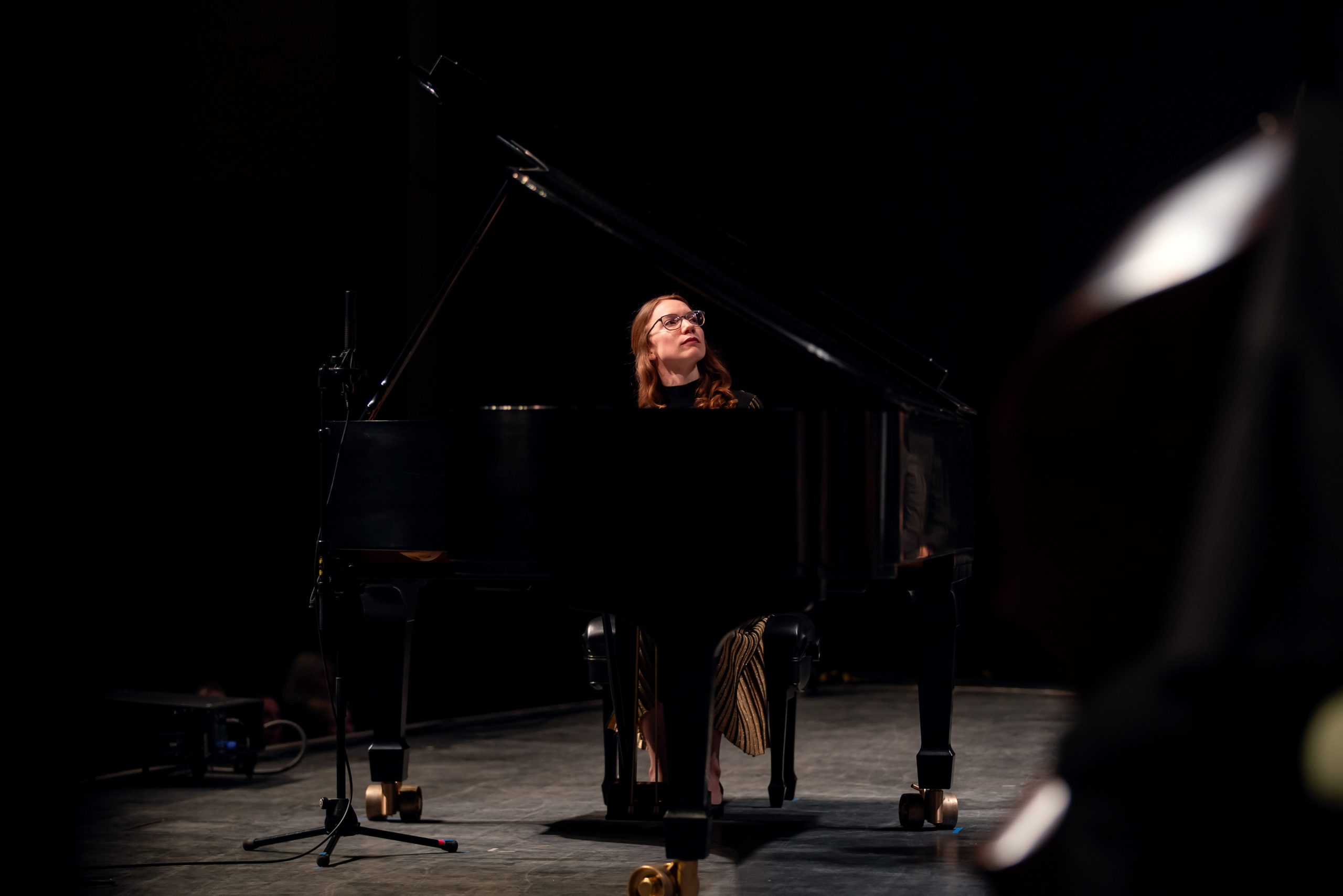 New World_Remai Modern A woman with long hair and glasses sits at a grand piano on a dimly lit stage. She looks upward thoughtfully as she plays. A microphone stand is positioned beside the piano, and a faint silhouette of another instrument appears in the foreground.