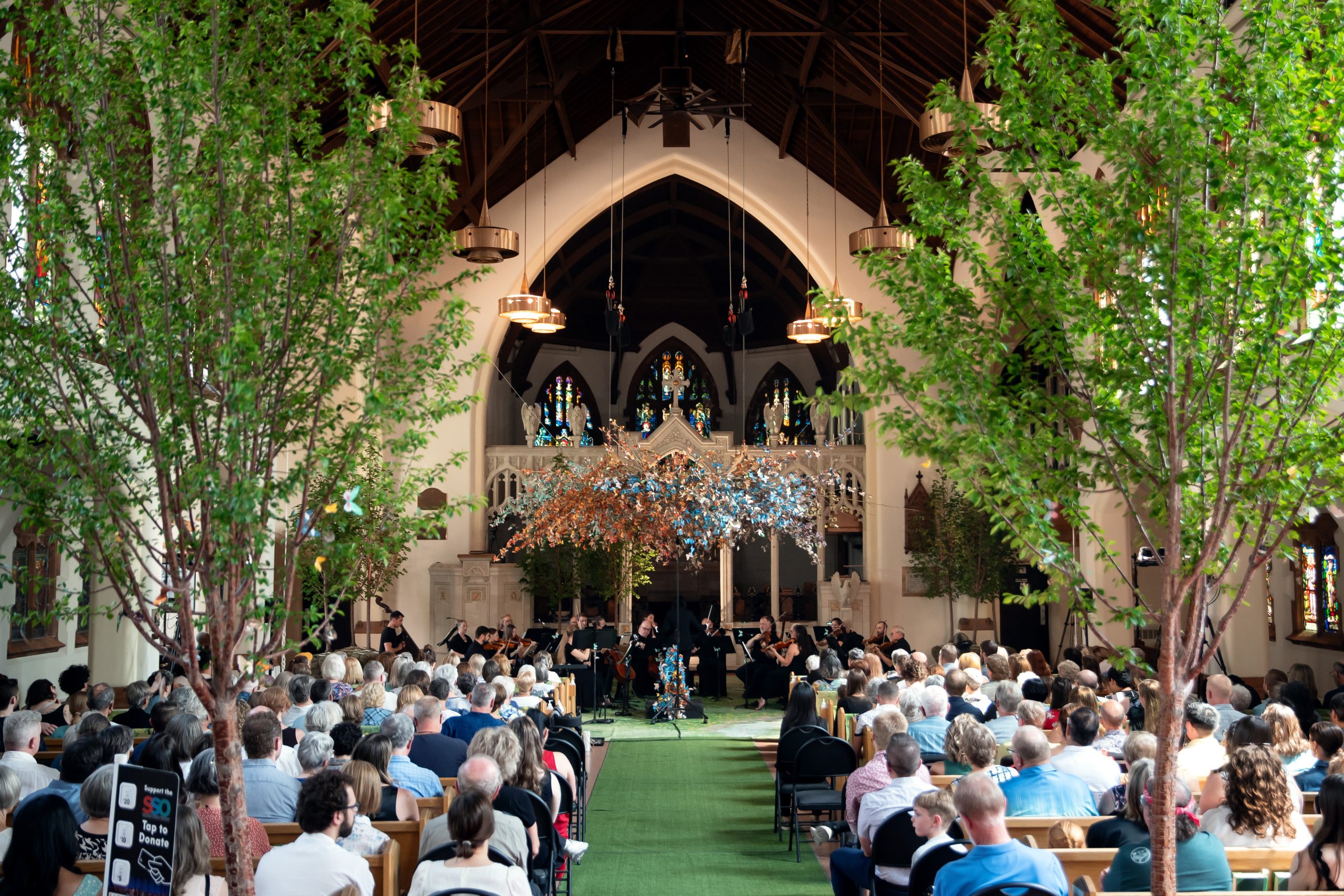 A large audience sits inside a church with high arched ceilings and stained glass windows, watching an orchestra perform at the front. Lush green trees and a large hanging installation of colourful branches decorate the aisle and stage, creating a natural, immersive atmosphere. The musicians are dressed in black, and warm light fills the space.