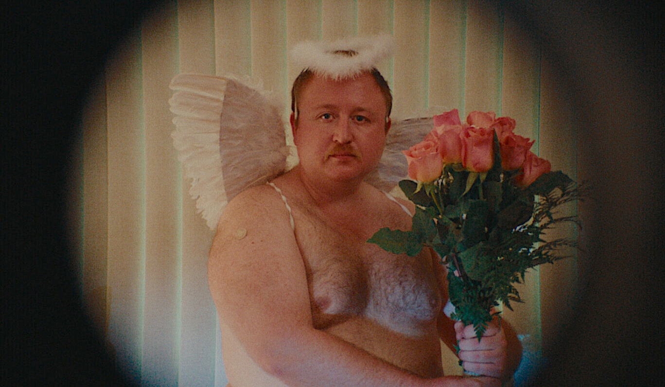 A man with a serious expression poses in front of vertical blinds wearing angel wings, a fluffy white halo headband, and white lingerie. He holds a bouquet of pink roses in front of his chest. The image has a soft, vintage look, with dark vignette framing.