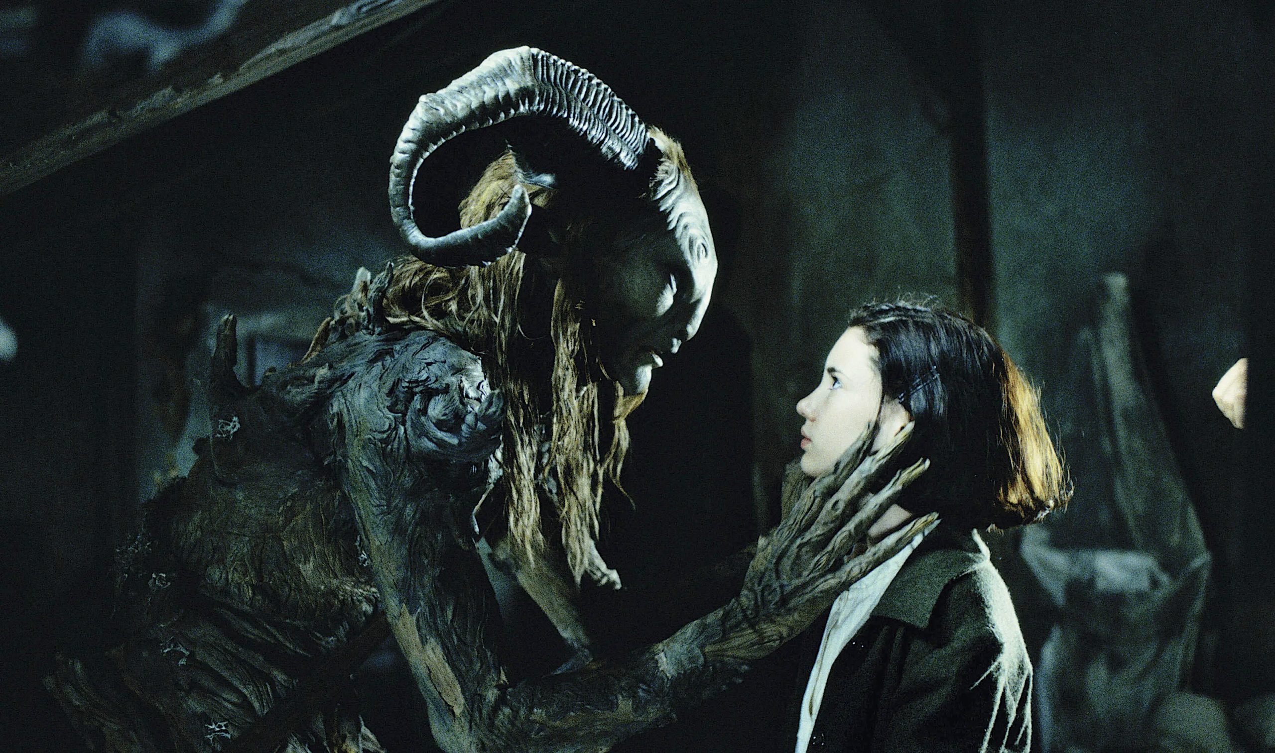 A fantastical creature with large curved horns, bark-like skin, and long hair gently holds the face of a young girl with dark hair. They gaze intently at each other in a dim, mysterious room with shadowy walls.