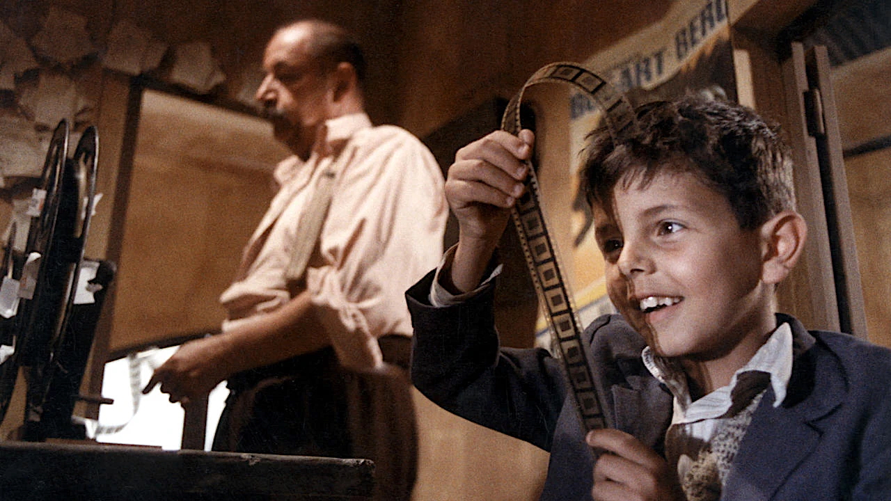 A young boy with tousled dark hair holds up a strip of film, smiling with excitement as he inspects the frames. He wears a collared shirt and a jacket. In the background, an older man in a beige shirt stands at a film editing machine, watching the boy fondly. The room is warmly lit and cluttered with film equipment and posters.