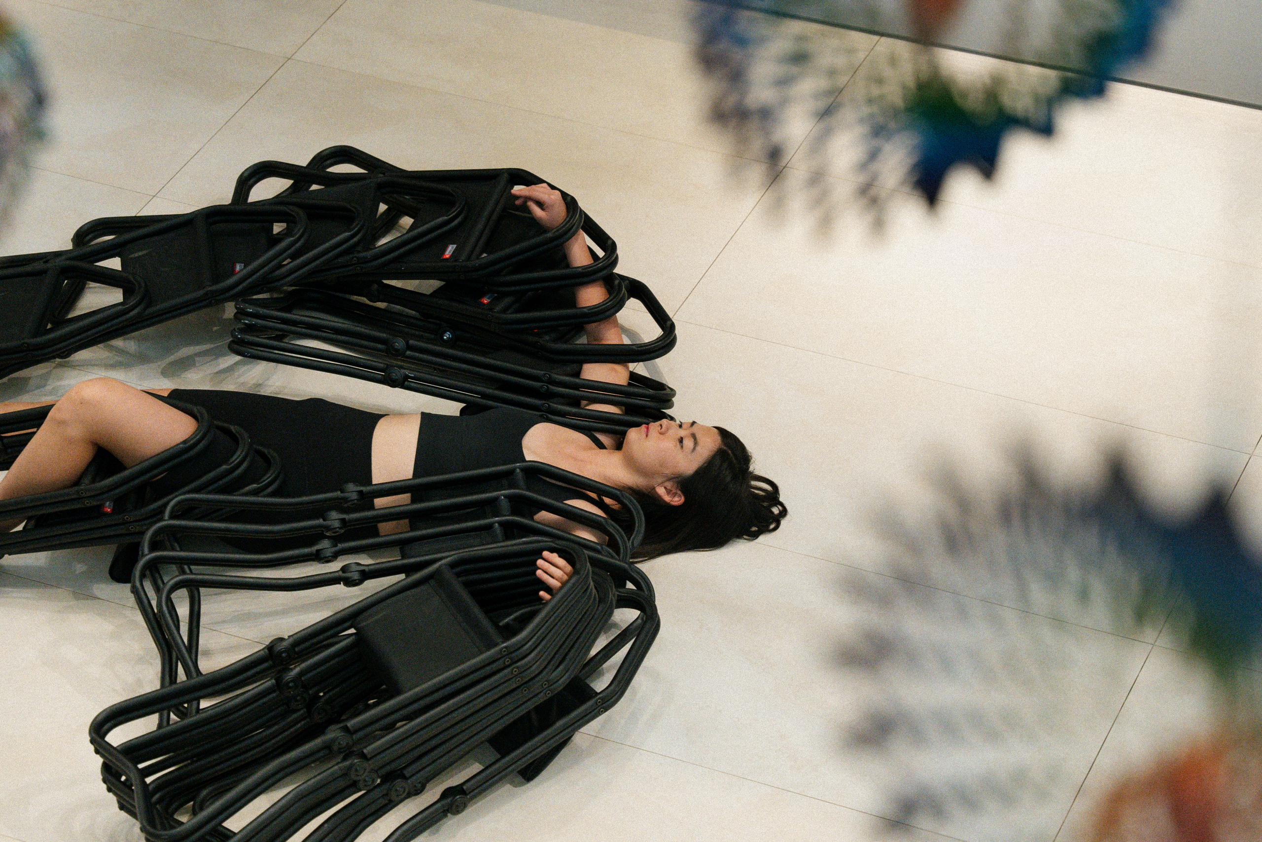 A woman in a black crop top and shorts lies on her back on a tiled floor with her eyes closed. Her arms and legs are intertwined with multiple stacked black folding chairs, almost encasing her body. The image is shot from above, with blurred colourful circular shapes partially framing the scene.