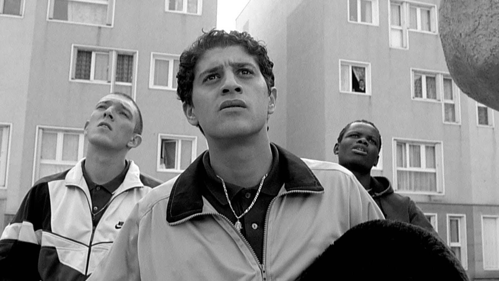 Three young men stand close together in front of an apartment building, looking upward with serious, concerned expressions. The black-and-white image highlights their track jackets and contrasting facial features.