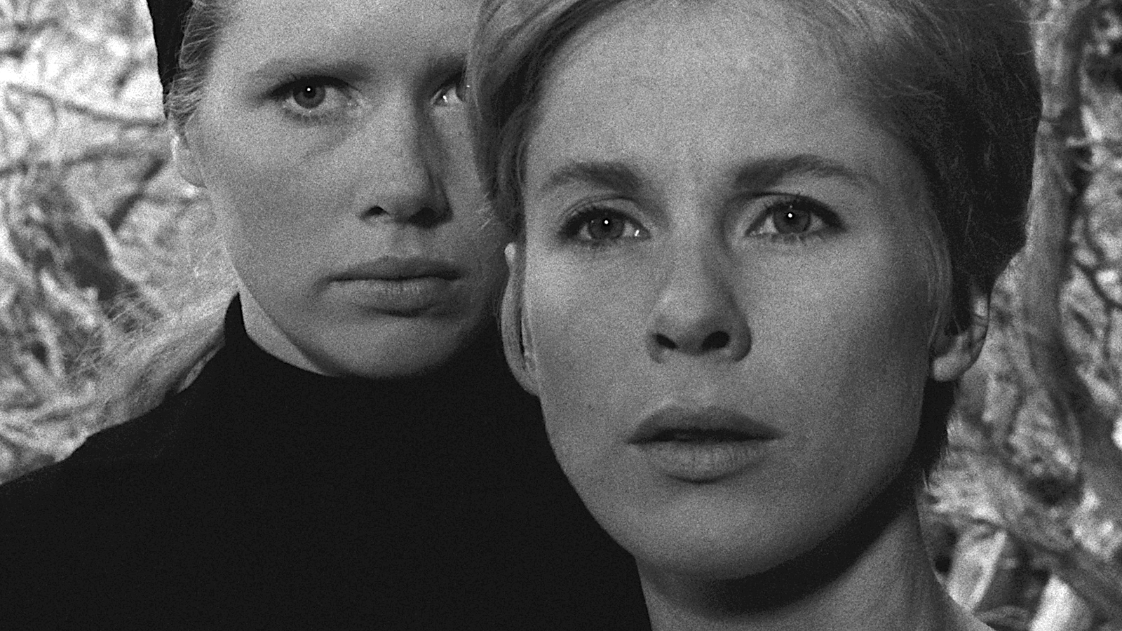 A black and white close-up of two women standing very close together. The woman in the foreground looks slightly to the right with an intent, serious expression, while the woman behind her gazes forward with an equally solemn look. Both wear dark clothing, and the background is softly out of focus, emphasizing their faces.