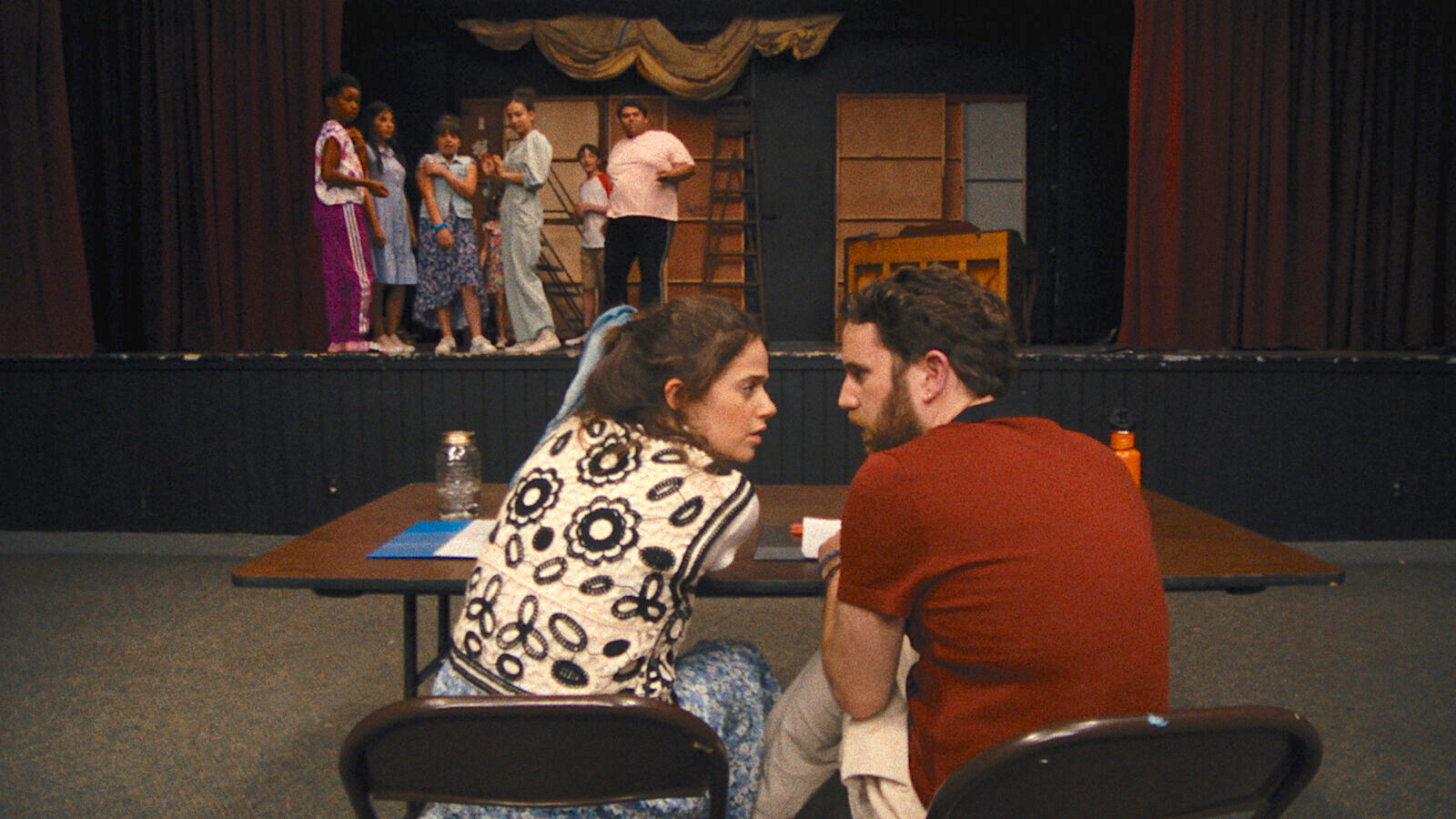 Two adults sit at a table facing a stage, leaning in to whisper to each other. On stage, a group of children in colourful clothes stand in a line, looking toward the adults. The stage is set with simple backdrops and a piano, suggesting a rehearsal in progress.