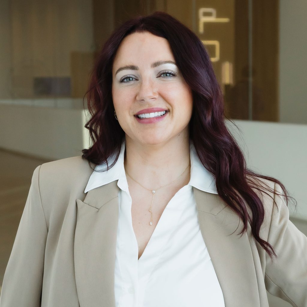 A person with long, dark auburn hair smiles at the camera. They are wearing a light-coloured blazer over a white blouse and a delicate necklace, standing indoors against a softly blurred modern interior background.