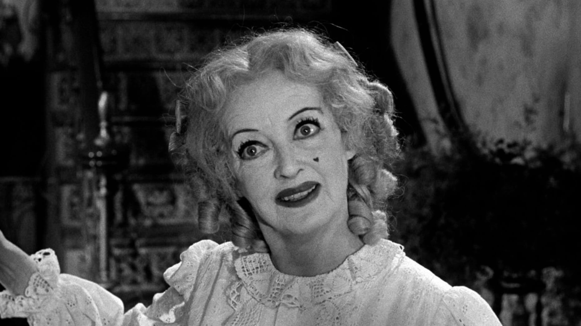 Black-and-white close-up of an older woman with tightly curled blonde hair in pigtails, wearing heavy makeup with exaggerated eyeliner, dark lipstick, and a beauty mark on her cheek. She smiles with wide, intense eyes and wears a frilly white dress, evoking a disturbing, doll-like appearance. The background is dimly lit and ornate.