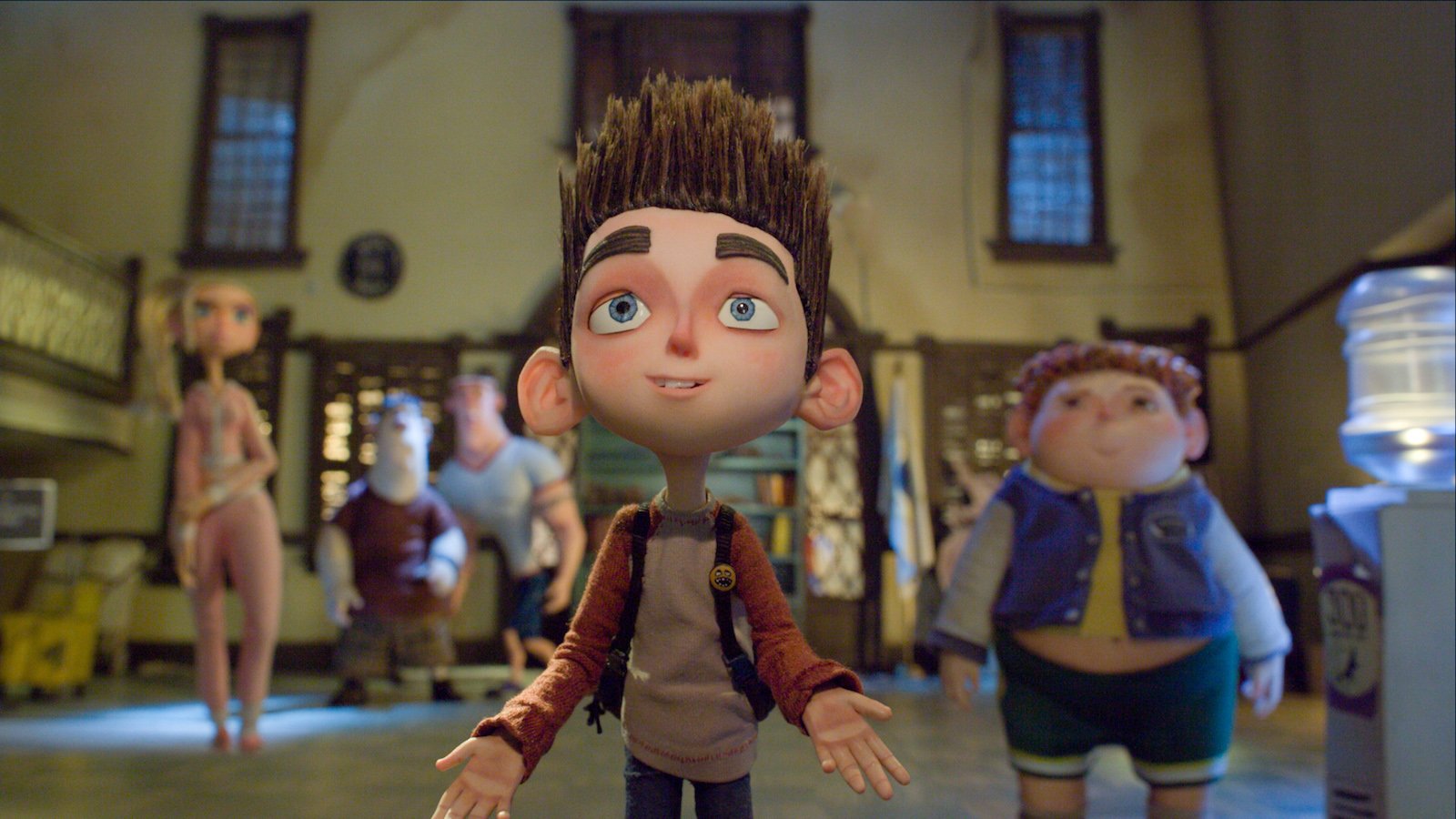A stylized stop-motion animated boy with spiky brown hair, large blue eyes, and oversized ears stands front and centre with his arms slightly open, wearing a brown and red hoodie. Behind him, several other animated characters stand in a dimly lit room, including a girl in pink, a stocky boy in green shorts, and two adult men. The background features tall windows and a water cooler to the right.