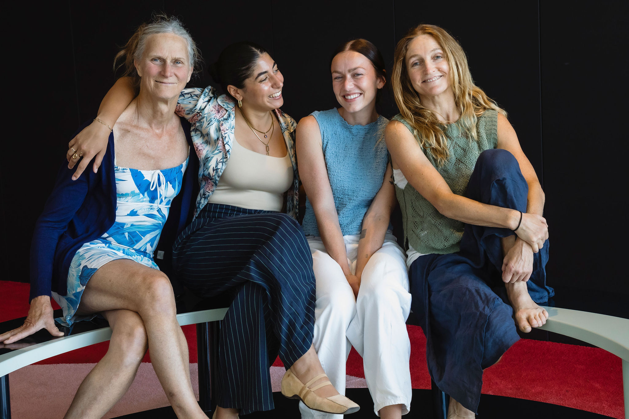 Four women sit closely together on a curved bench, smiling warmly. Each woman wears a different summery outfit—ranging from dresses to pants and tops—with relaxed, natural expressions. The background is dark, contrasting with the bright, casual clothing and the soft red carpet beneath them. Their body language conveys connection and camaraderie.
